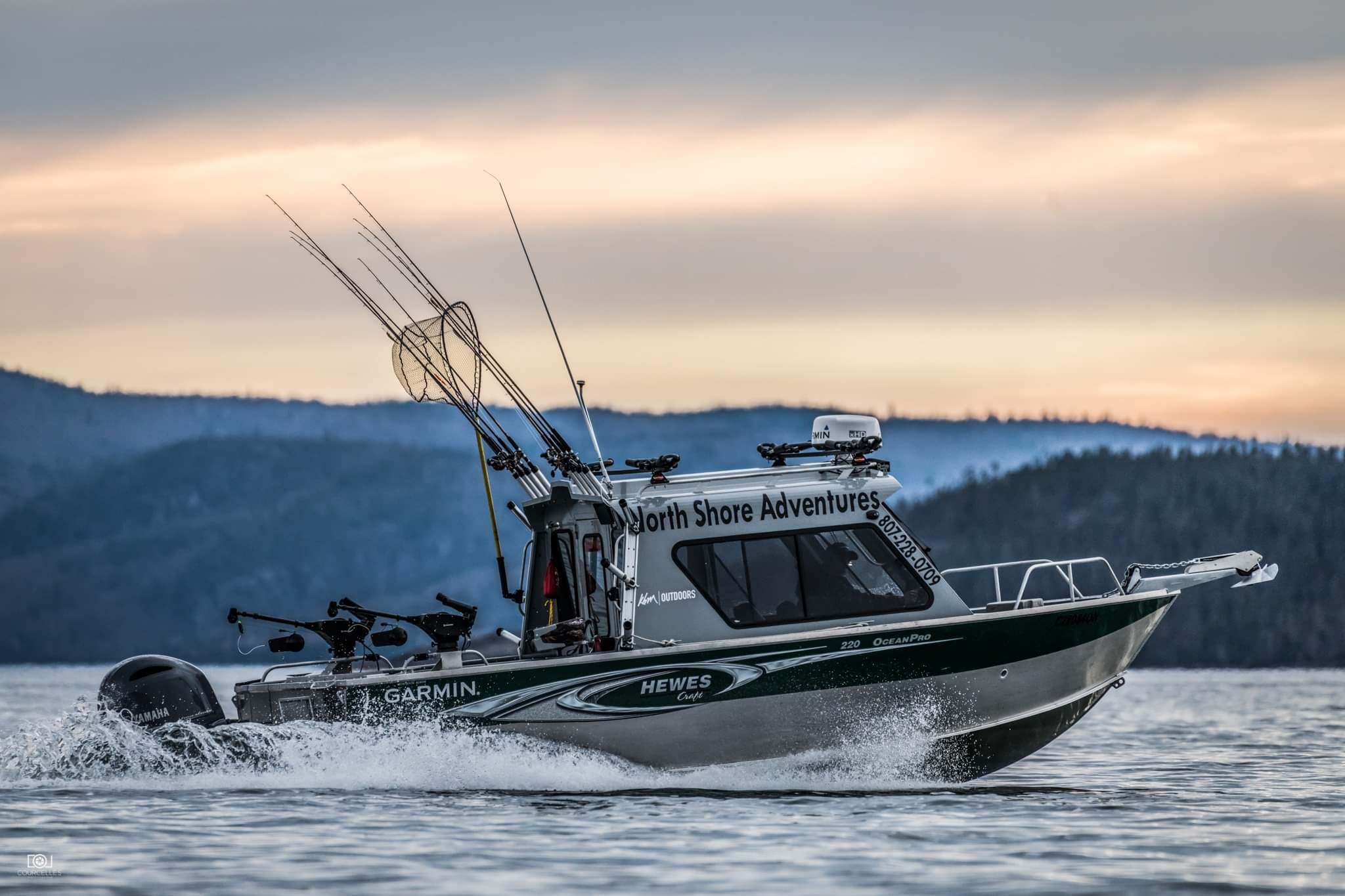 a fishing boat with nets and rods in the back zips across the water of Lake Superior, with misty forested hills in the background.