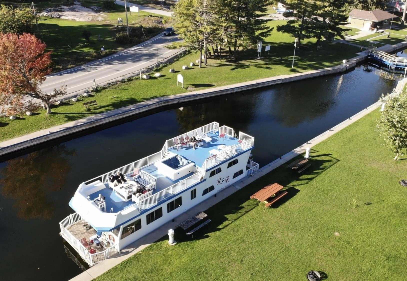 A houseboat sits in a lock along the Trent-Severn Waterway. The locks are surrounded by lush green grass, trees and park space and the weather is sunny.