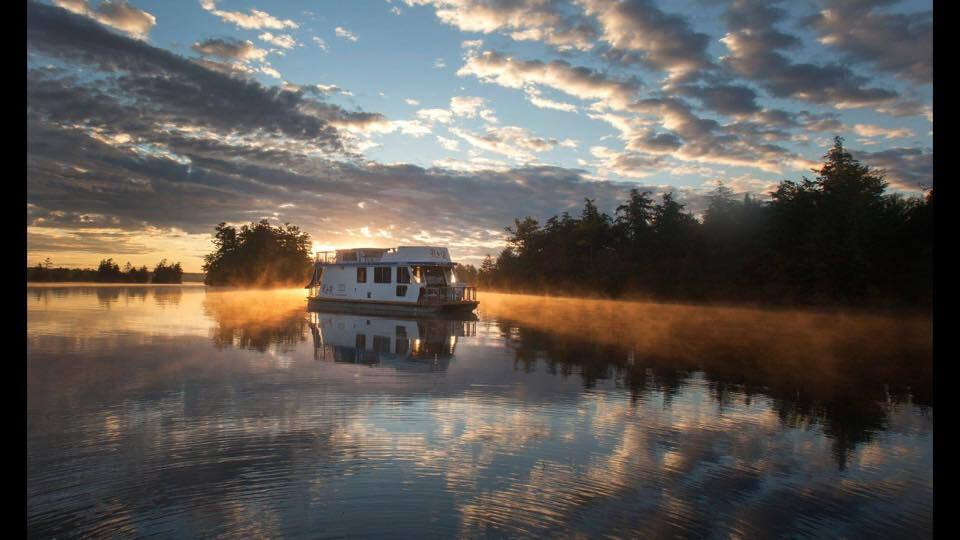 A houseboat sails on calm glassy waters under an orange sunset along the Trent-Severn Waterway