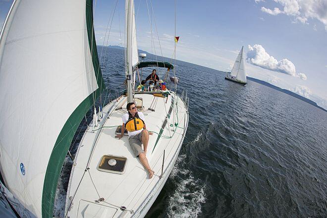 a man wearing sunglasses and a life preserver smiles as he reclines on the deck of a sailboat in the sun, looking out over the wavy blue of Lake Superior.