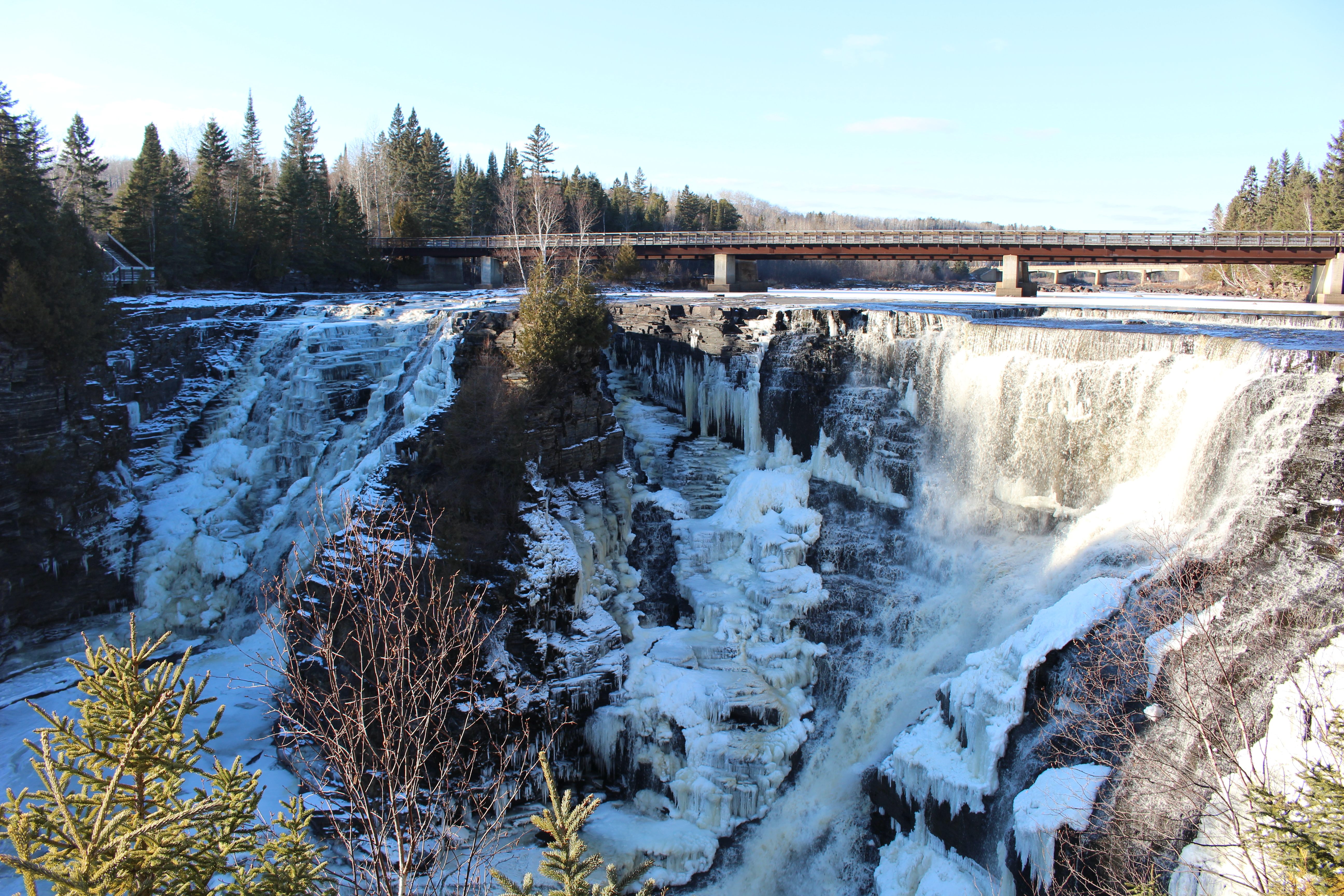 Kakabeka Falls in the winter, frozen into a tall jagged ice cliff surrounded by forest.