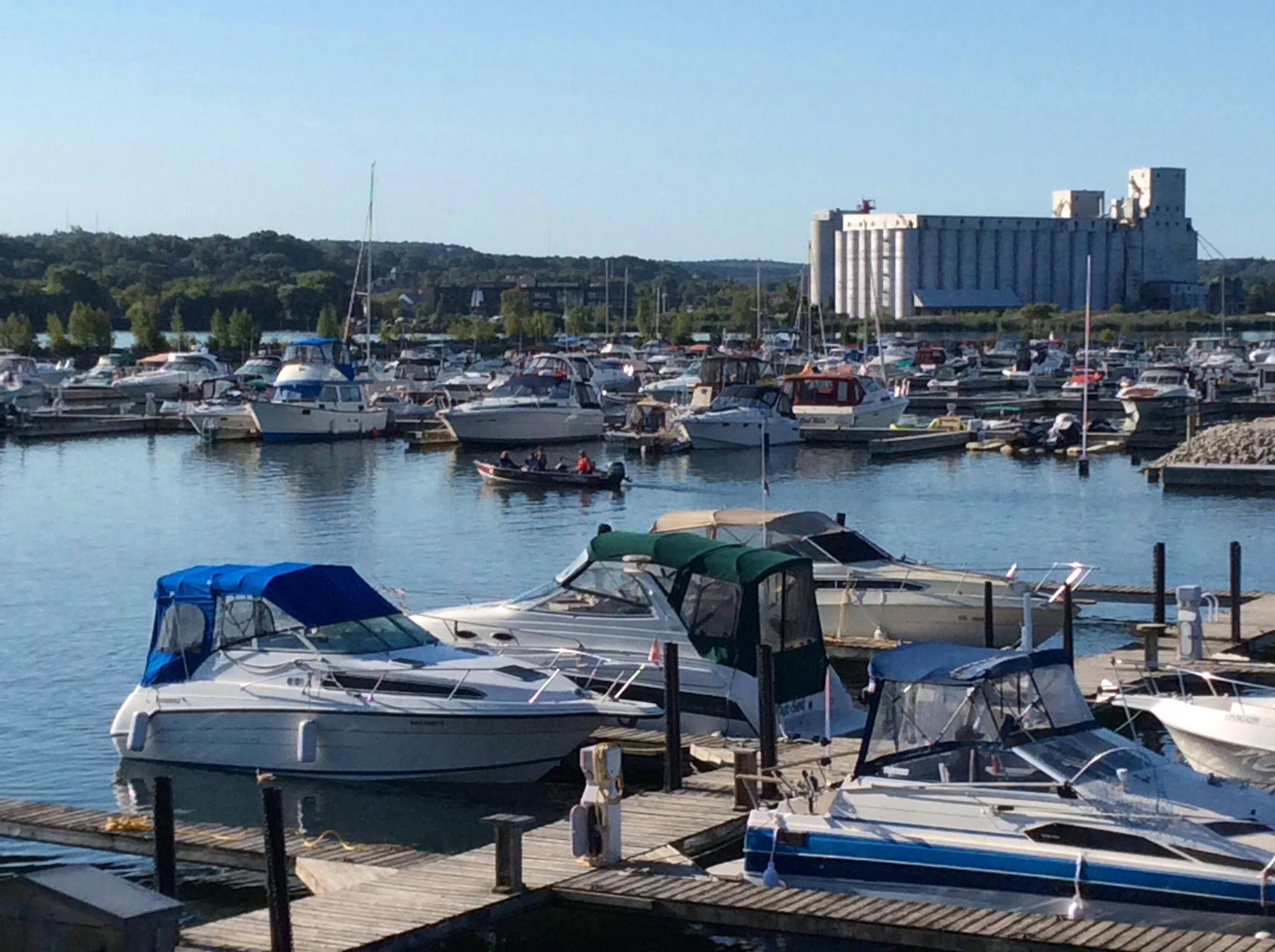 Georgian Shores Marina; a large, busy marina with many small boats docked along its slips. The still water and clear sky are blue and there are green forested hills beyond the marina.