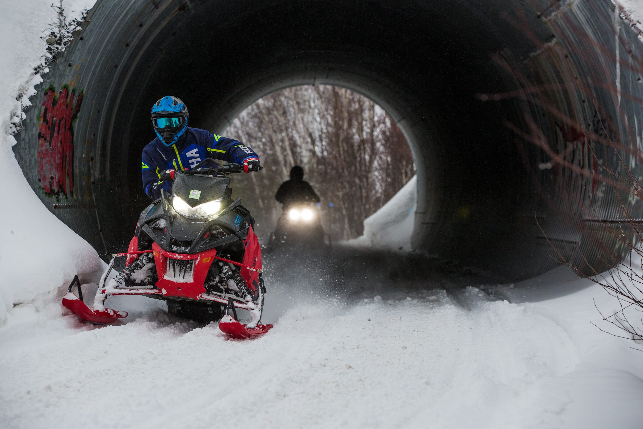 two snowmobilers ride through a dark narrow tunnel over a snowy trail.