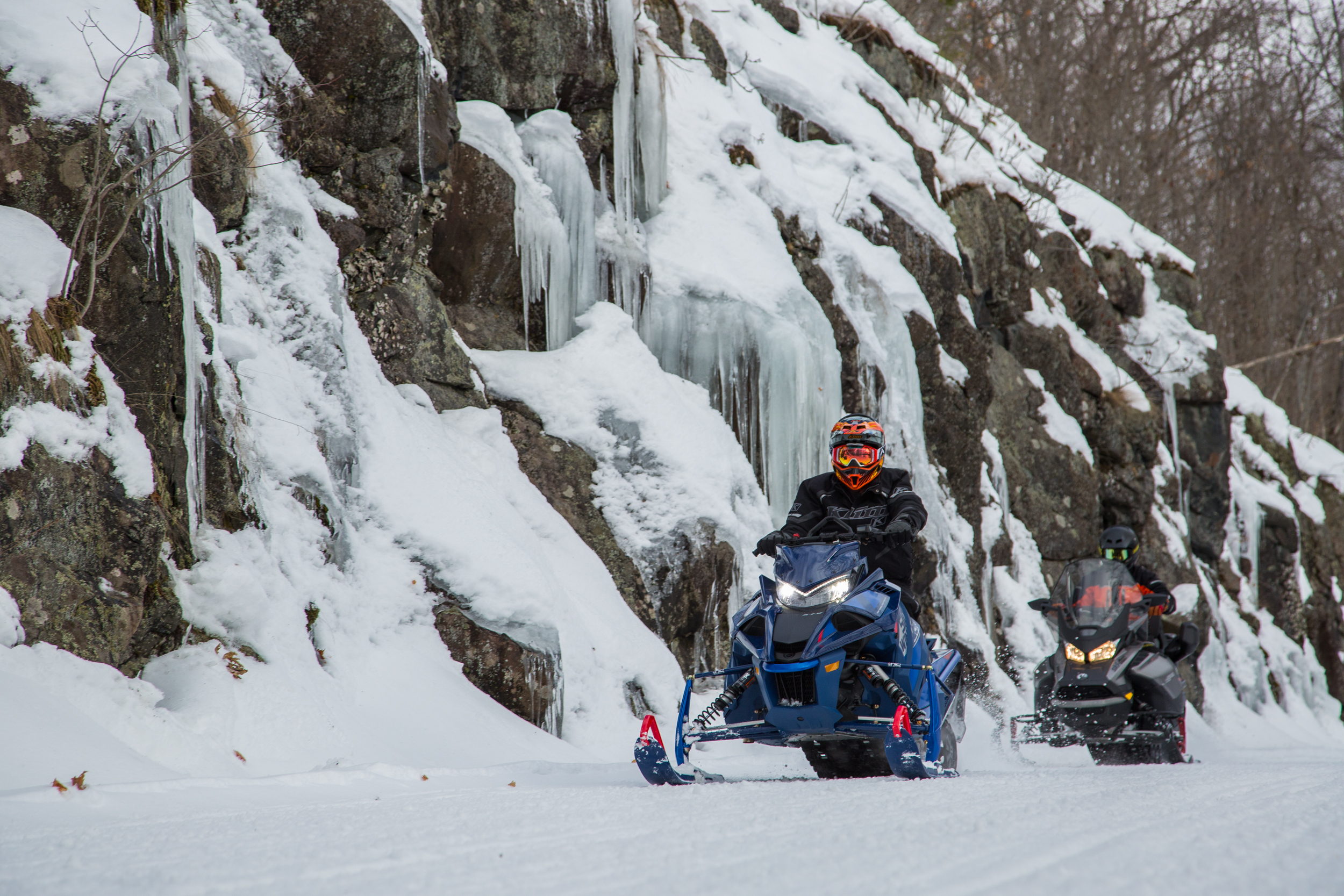 snowmobilers ride next to a tall rocky cliff covered in snow and ice.