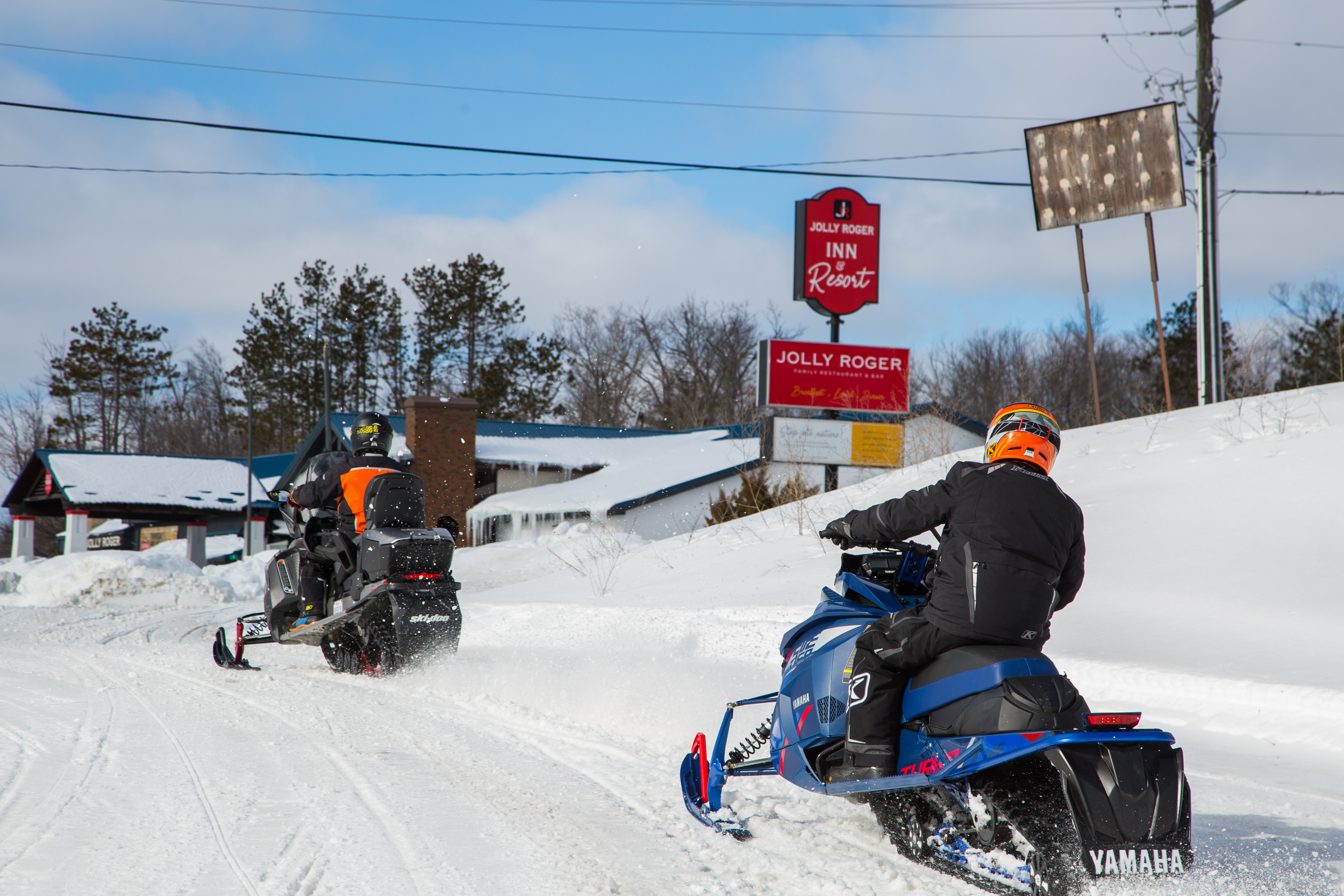 two snowmobilers ride up a snowy trail toward the Jolly Roger motel.