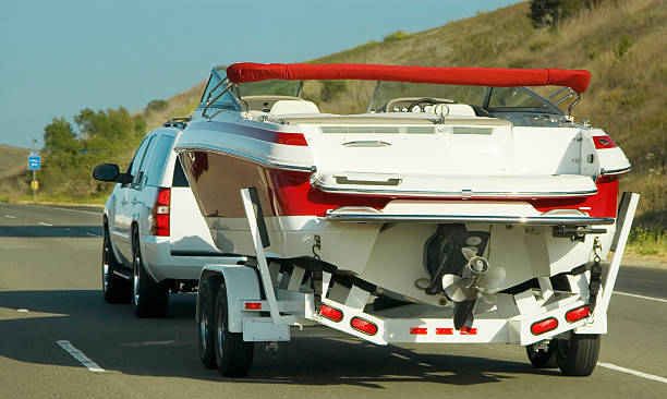 A truck tows a red and white powerboat on a trailer along a paved highway surrounded by green trees, hills and brush.