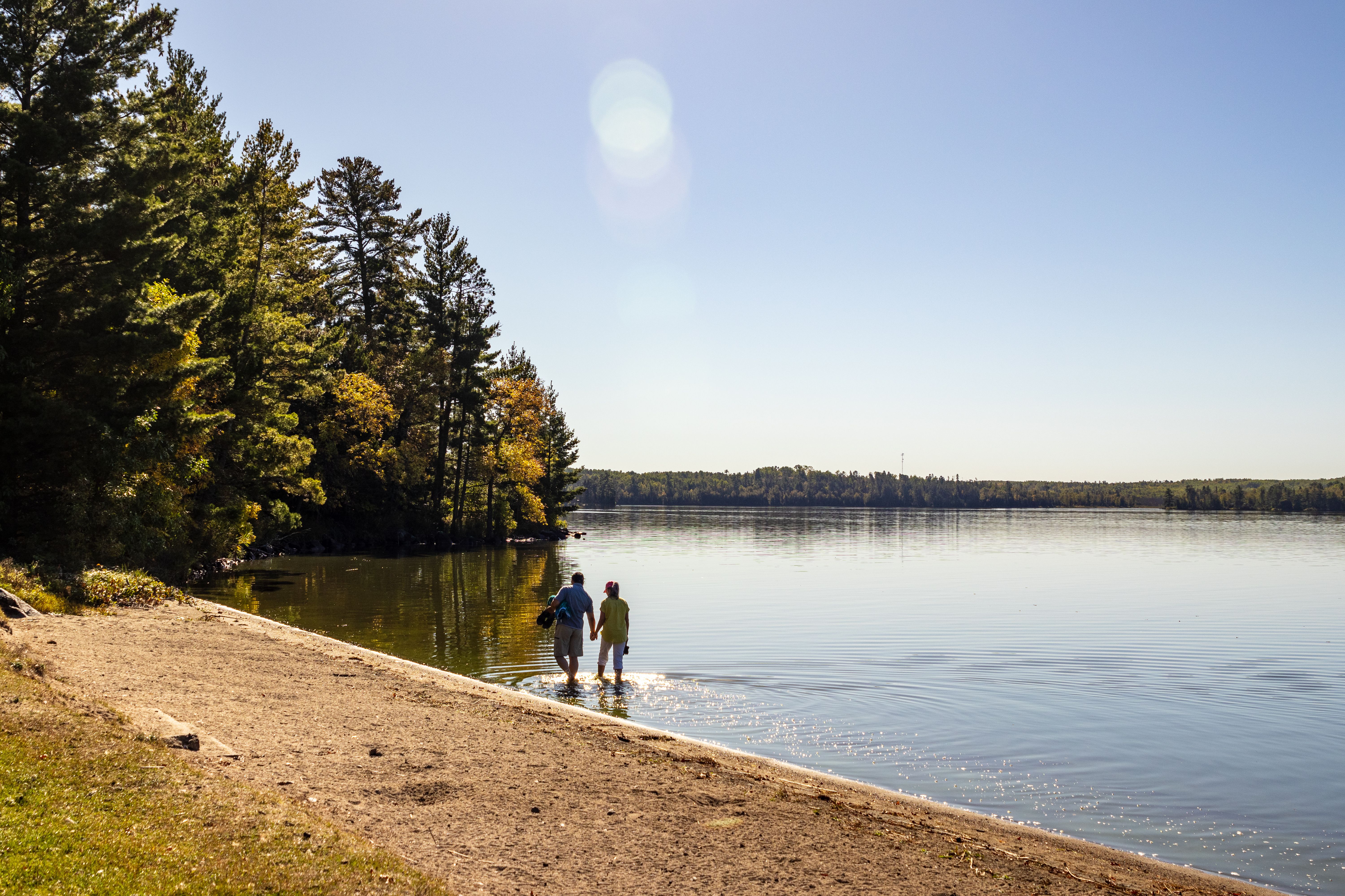 A couple walks hand in hand, wading along the shoreline at Caliper Lake Provincial Park on a clear sunny summer day.