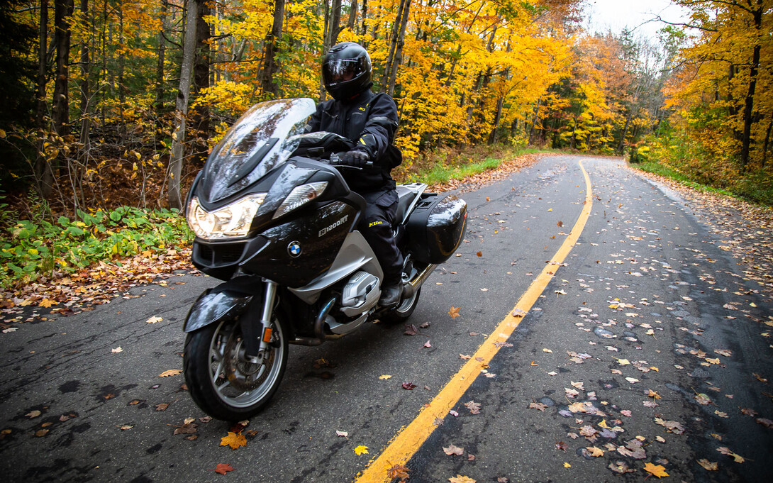 a motorcylist drives down a narrow, smooth paved road through a forest lit up by vibrant yellow autumn leaves.