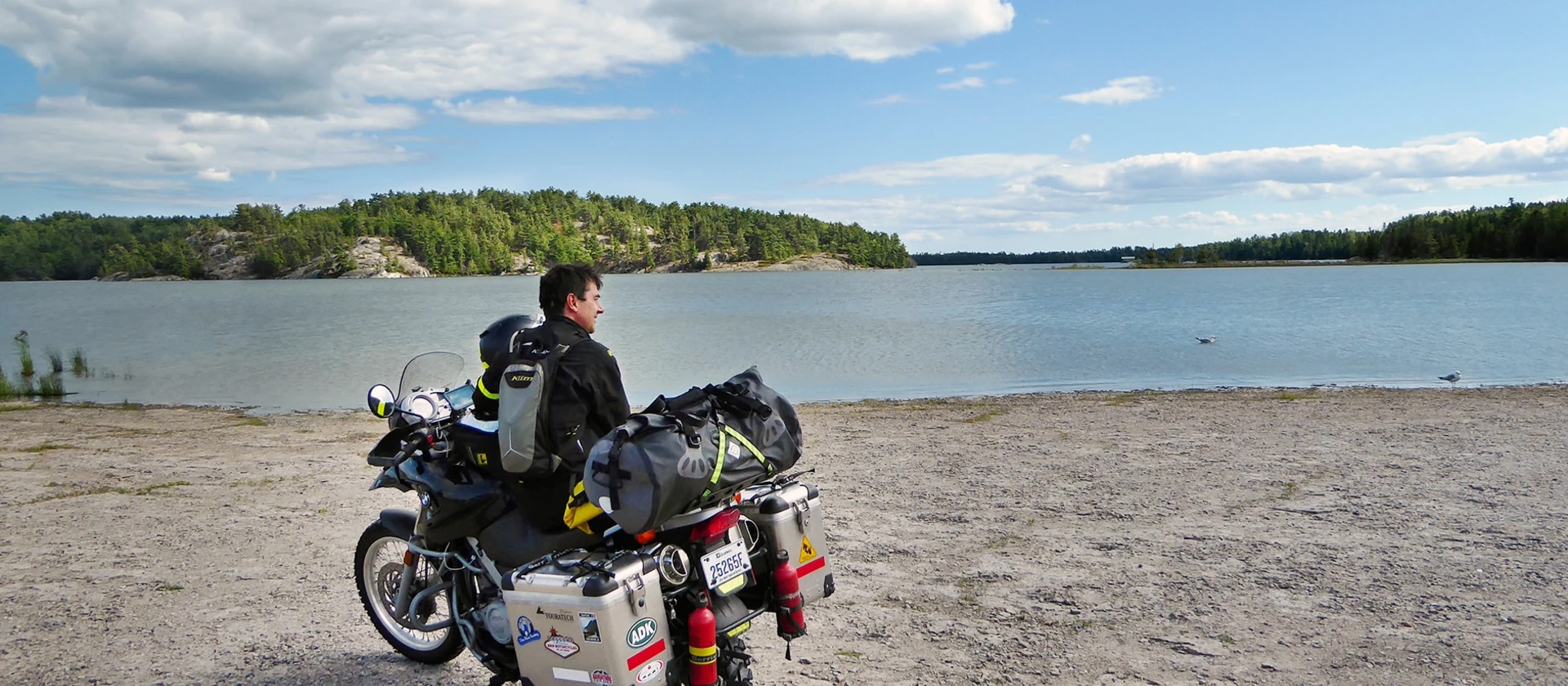 A motorcyclist parked and resting on a beach next to a large blue lake on a summer day.