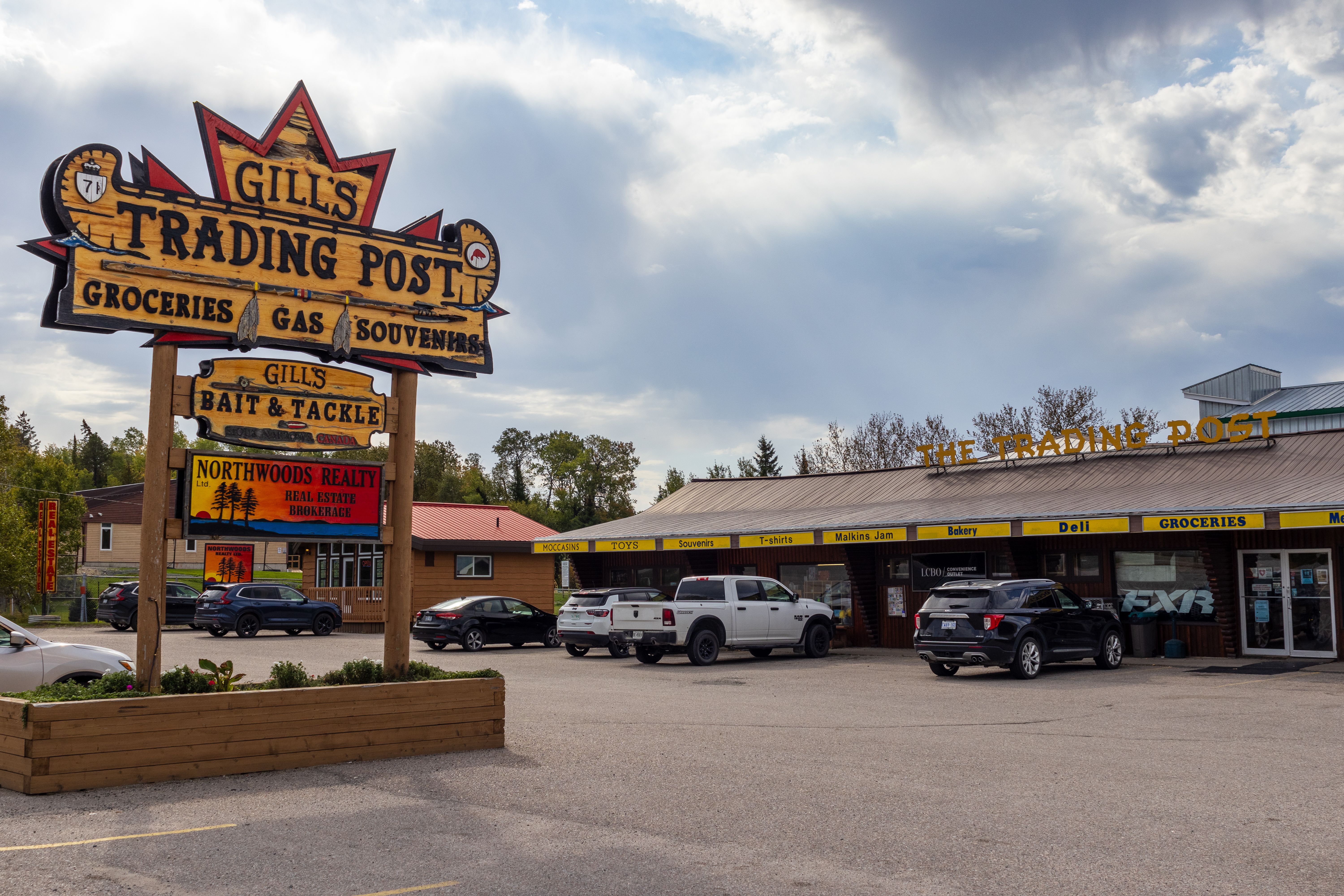 Gill's Trading Post; a long wooden shop building with an ornate wooden sign shaped like a maple leaf advertising the business's groceries, gas and souvenirs. 