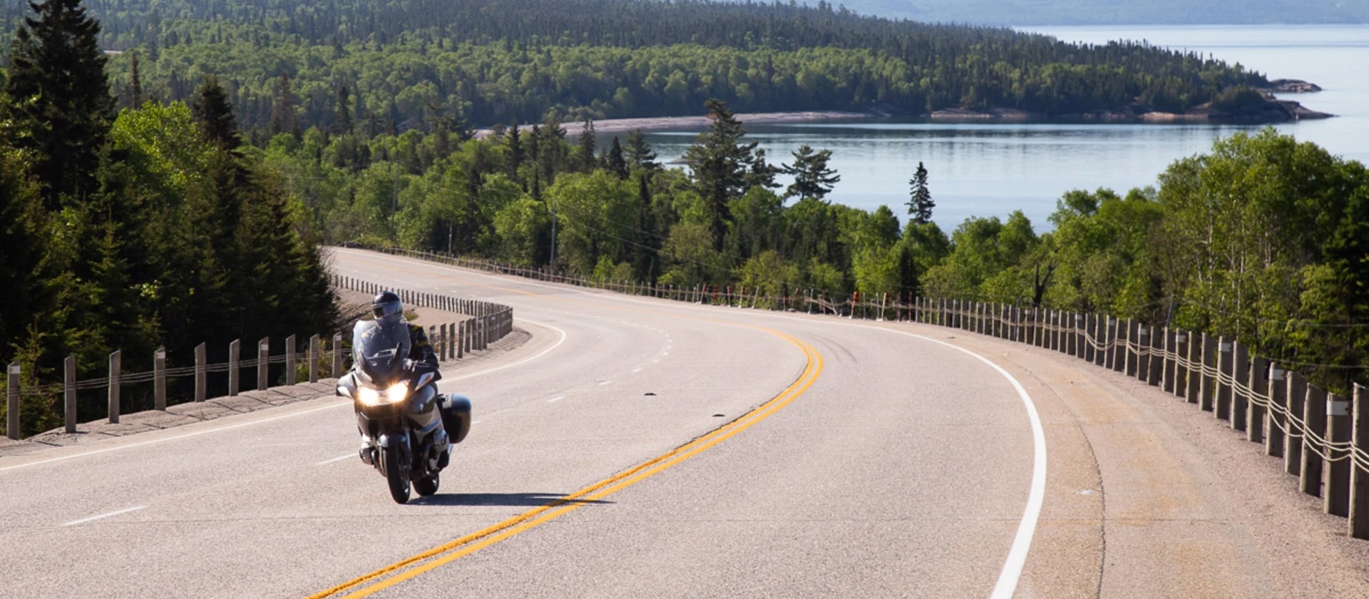 a motorcyclist rides along a smooth paved highway along the edge of Lake Superior, surrounded by thick green boreal forest.