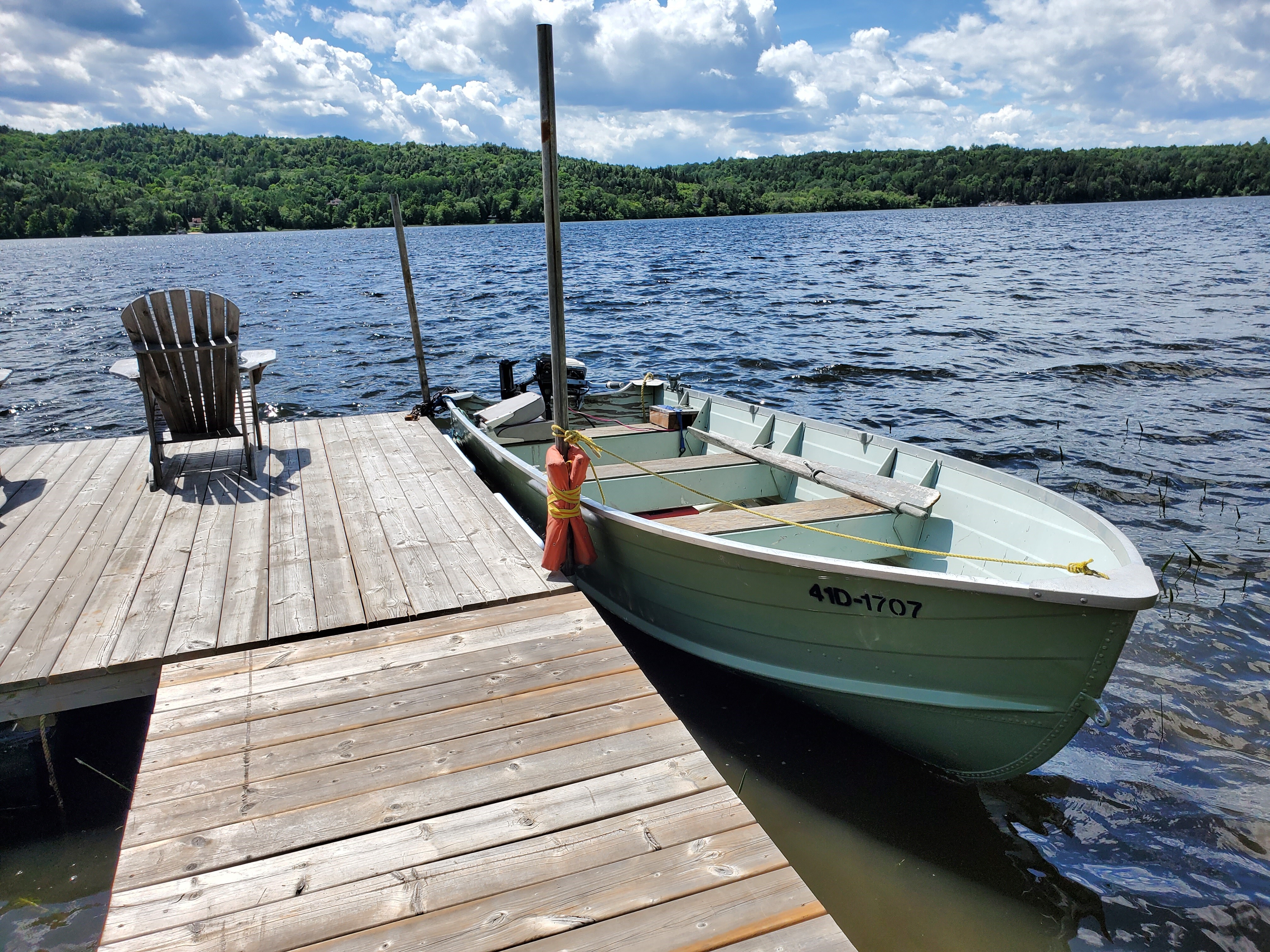 a small boat along a wooden dock, floating on a peaceful blue lake with forested shoreline. 