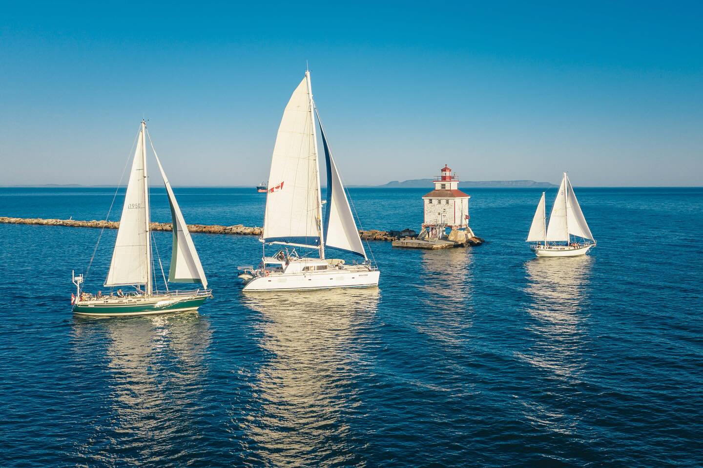 3 sailboats float on glassy water under a clear blue sky next to a small lighthouse in Lake Superior.