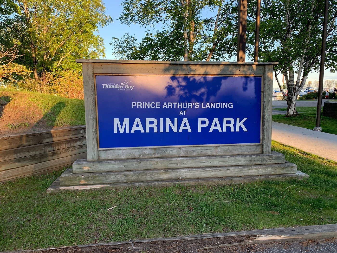 The wooden and blue Thunder Bay Marina Park sign next to the waterfront, surrounded in green trees and grass.