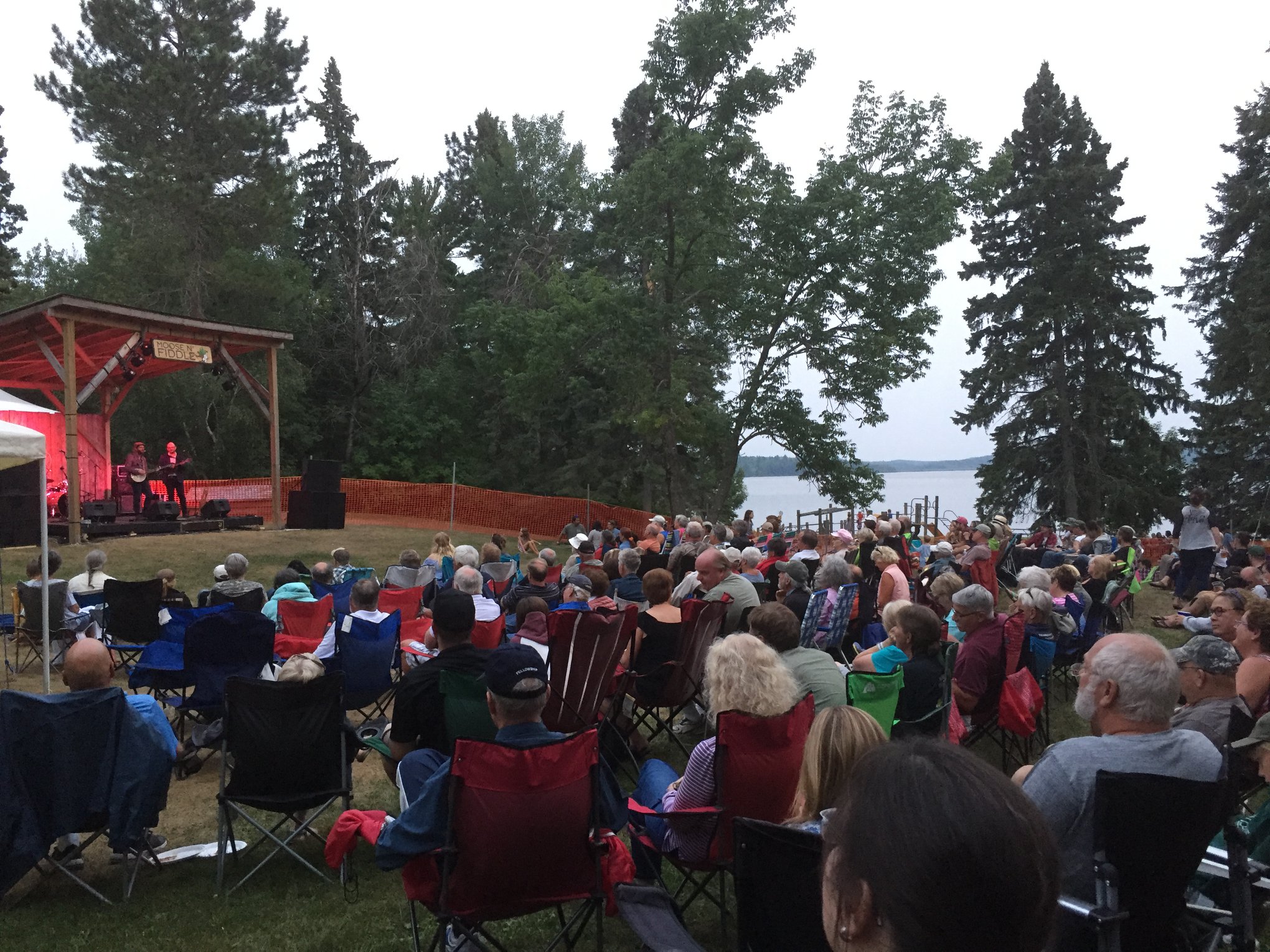 A crowd of people in foldable chairs sit next to a lake on a summer evening watching a band play on and outdoor stage labelled "Moose and Fiddle".