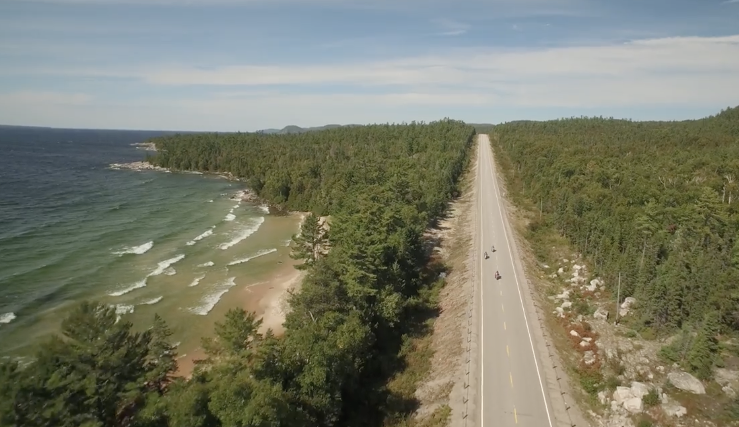 highway 17 stretching off into the horizon through green boreal forest, with 3 motorcyclists driving along it. A turquoise Lake Superior laps waves against the shore next to the highway.