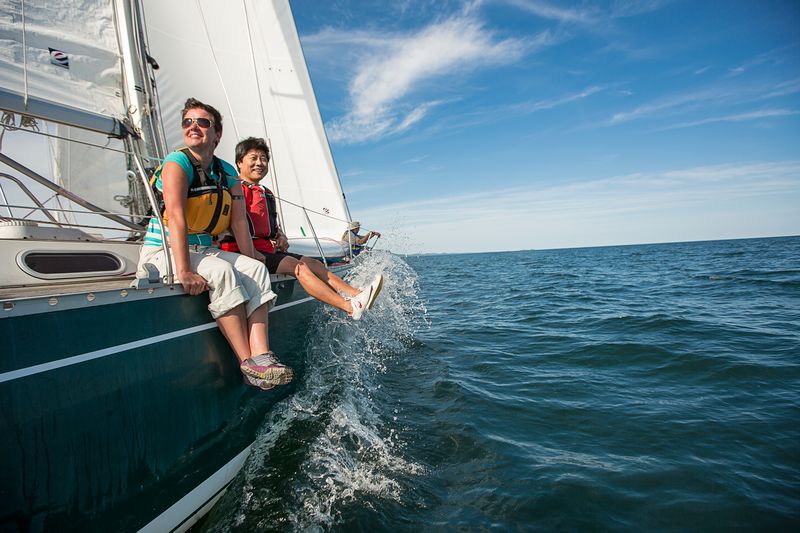 Two smiling people dangle their feet over the edge of a sailboat as the water from Lake Superior splashes up around their feet. 