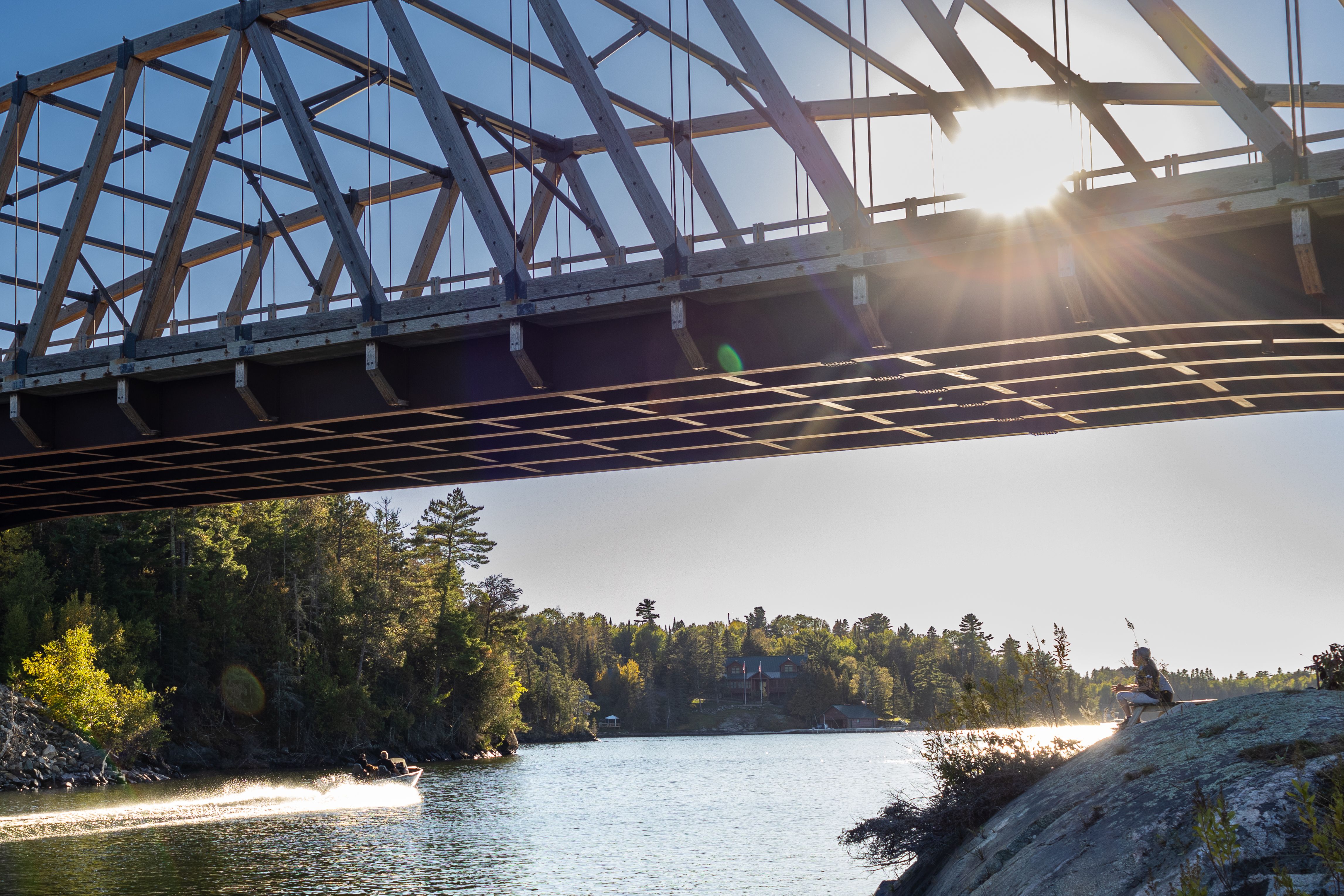 The large wooden heritage bridge over the river at Sioux Narrows-Nestor Falls on a summer day. A person sits on the rocks under the bridge, looking at a boat zoom by on the shining water. 