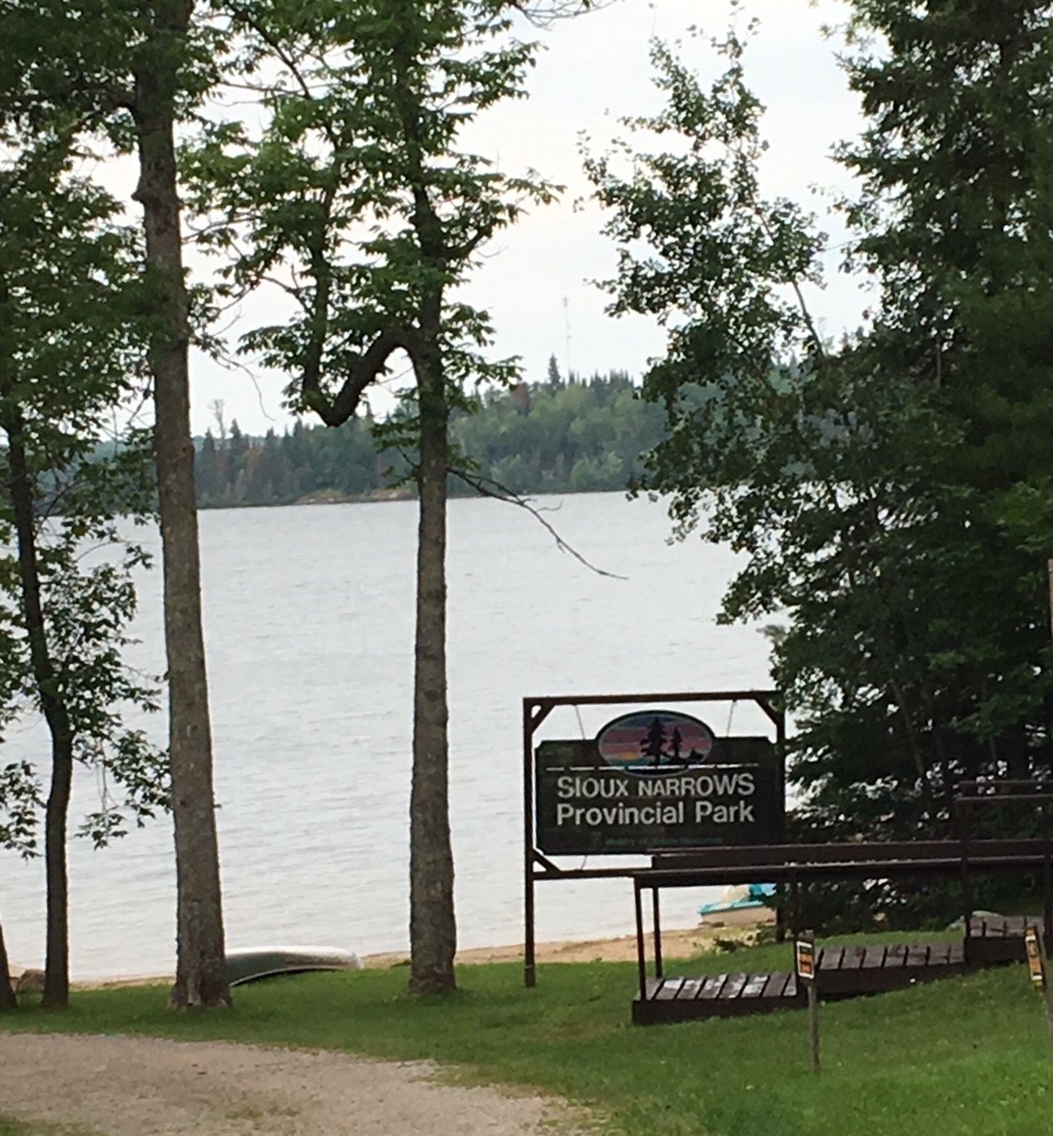 The Sioux Narrows Provincial Park sign in front of a lake surrounded by trees, next to a dirt path. 