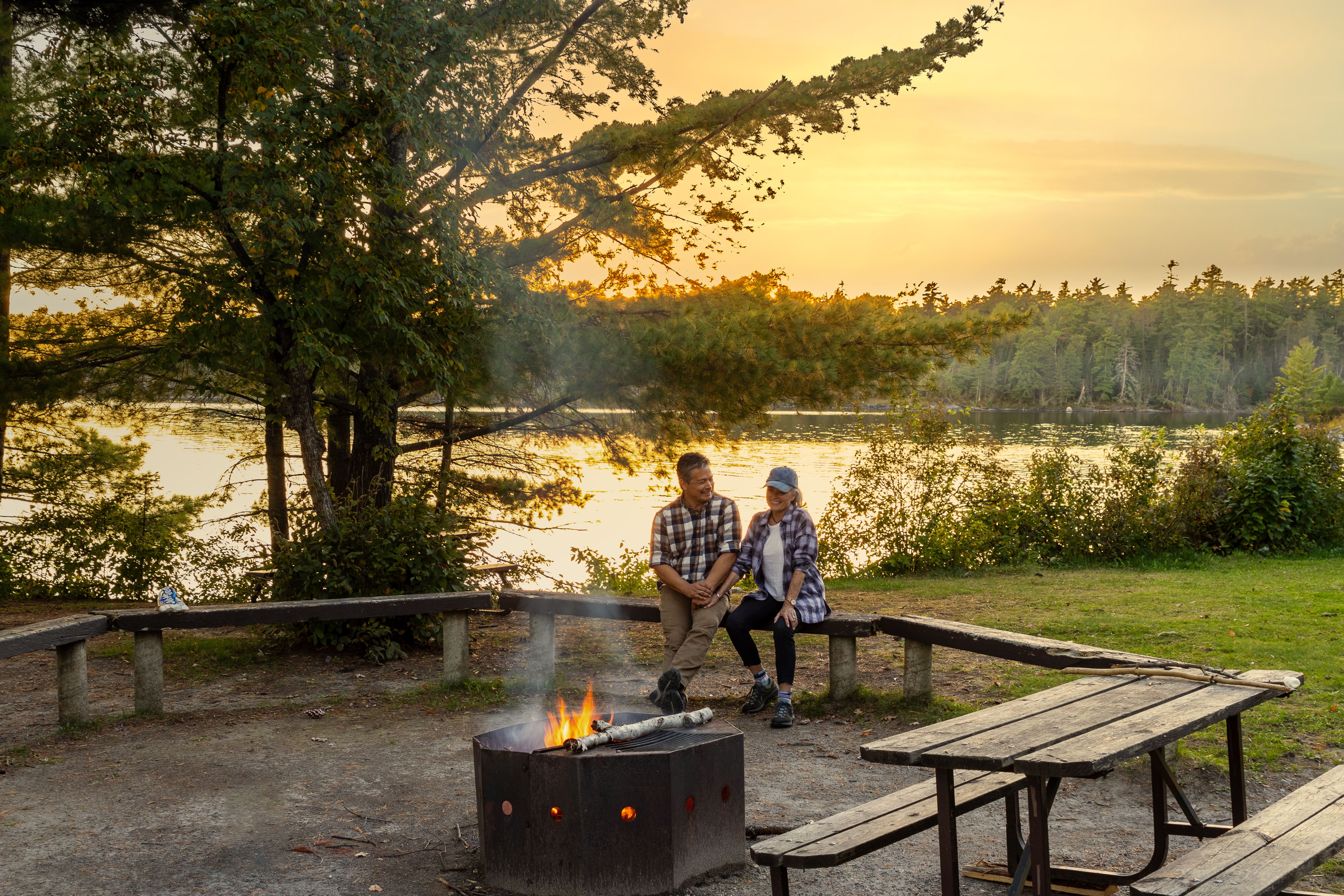 Sioux Narrows Provincial Park; a couple chats over their campfire at sunset in front of a lake surrounded by forest.