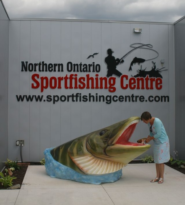 a woman looks into the mouth of a very large colourized statue of a fish rising out of the floor under the Northern Ontario Sportfishing Centre sign. 