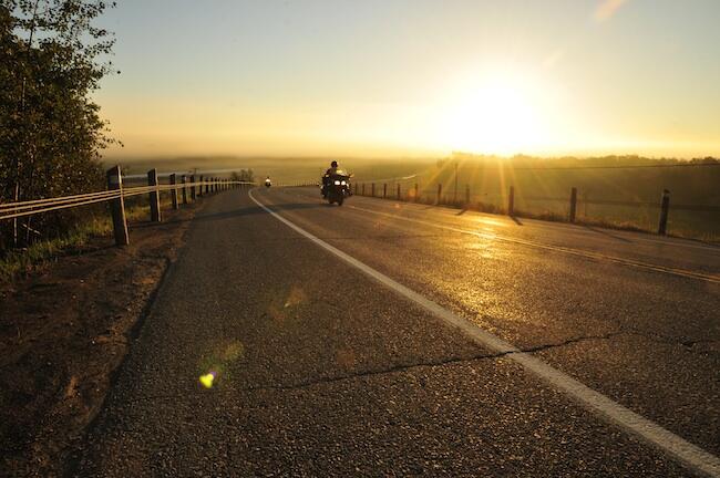 A motorcyclist rides along a flat, smooth highway through green pastures, lit up by a golden summer sunset under a perfectly clear sky.