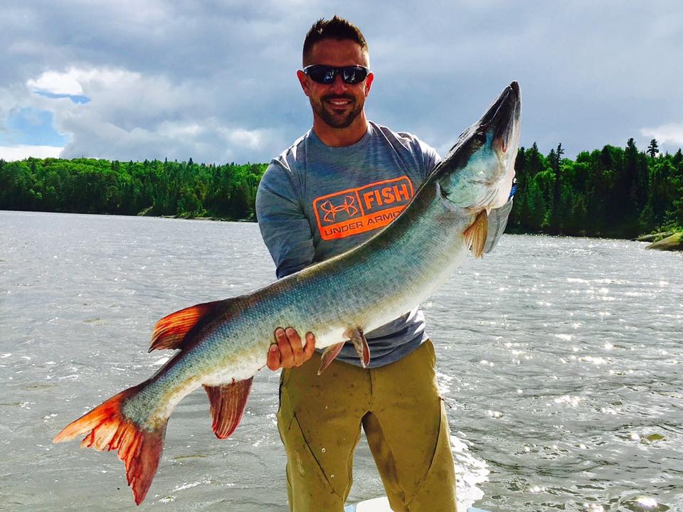 A smiling man holds up a large muskie about two-thirds the length of his body while standing on a boat on a shining lake. 