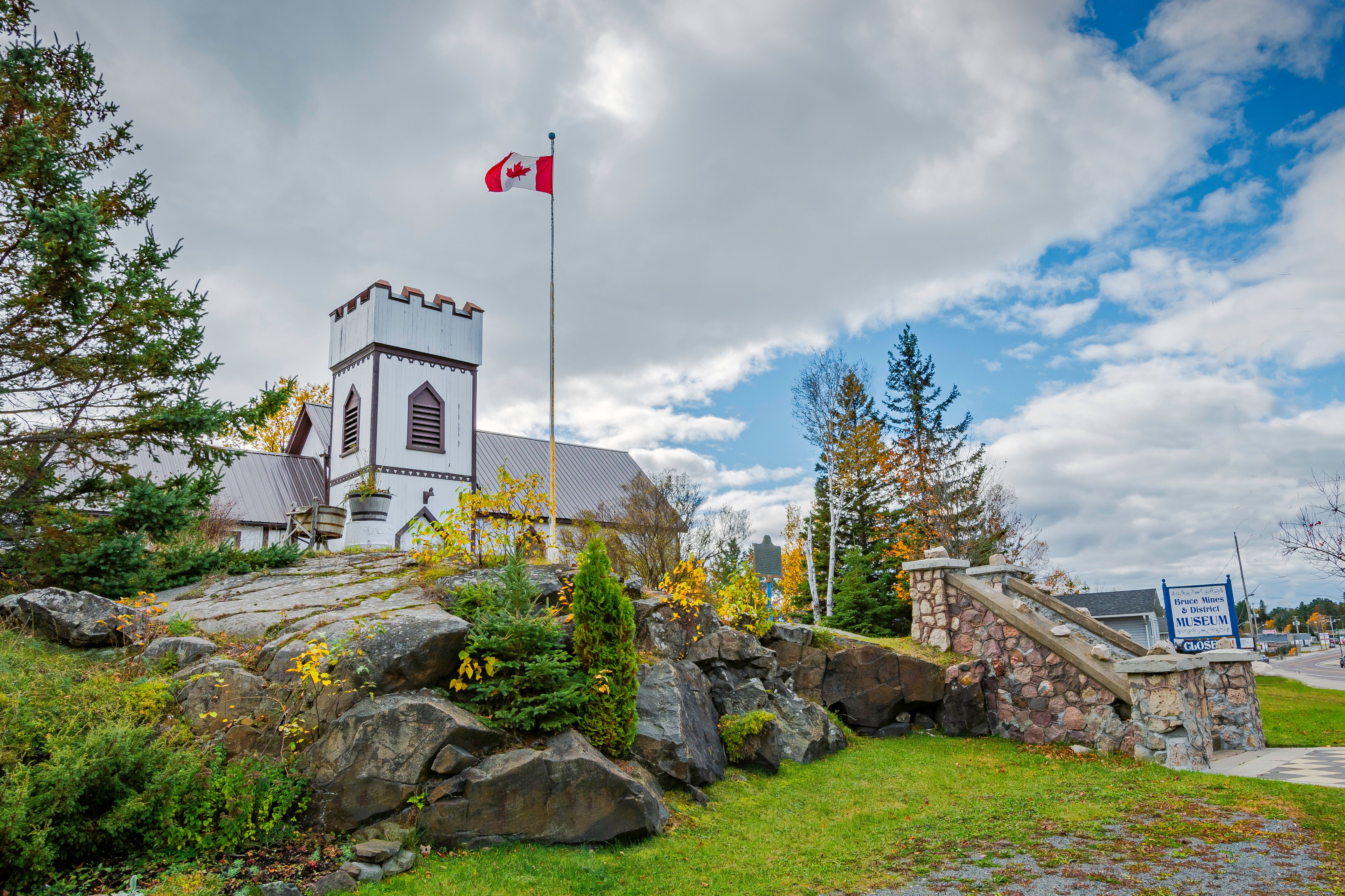 The Bruce Mines Travel Information Centre; a stately and interesting wooden castle-shaped building with stone stairs, green lawns and boulders outside.