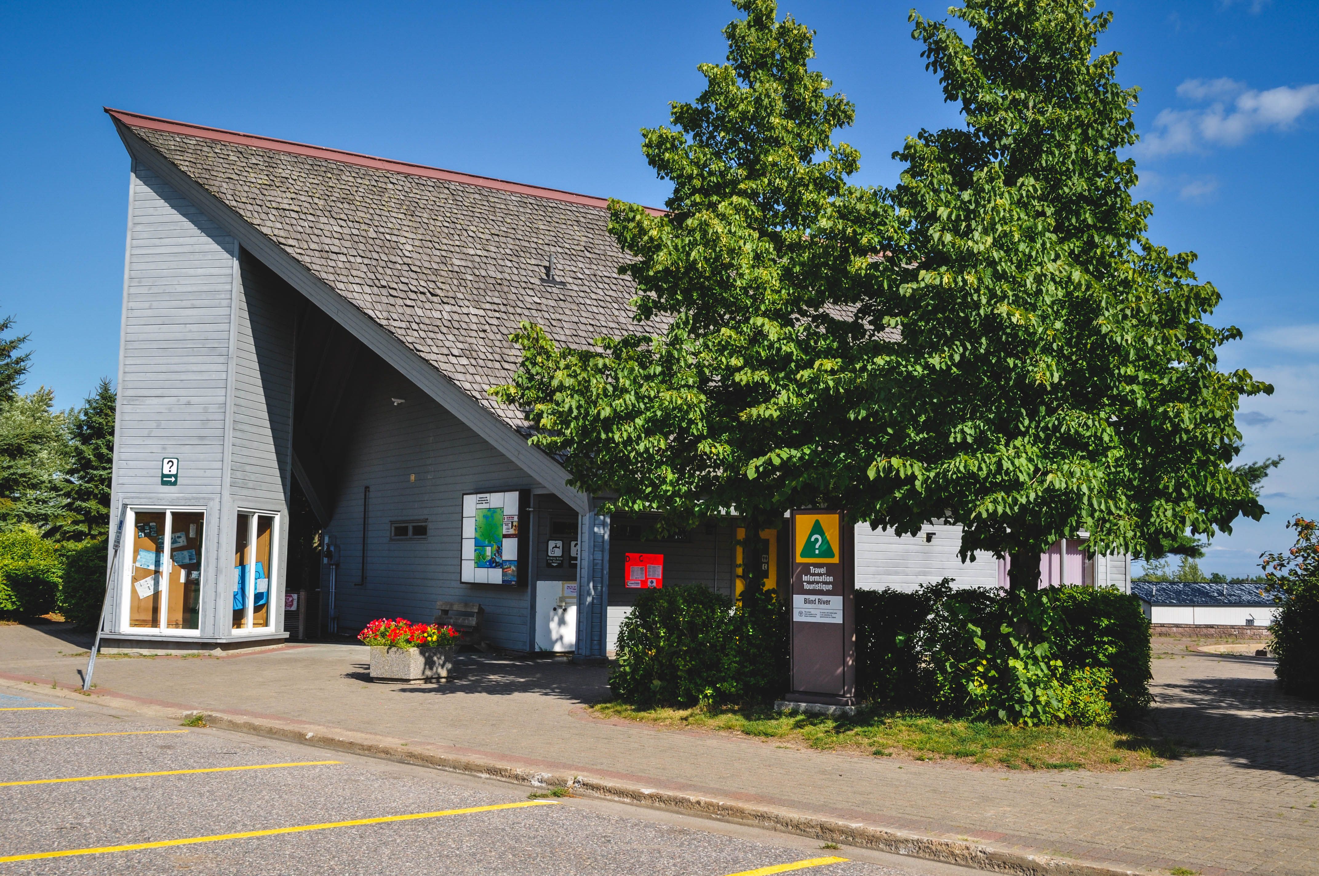 The Blind River Travel Information Centre; a tidy grey, modern-looking building with lush green trees outside.