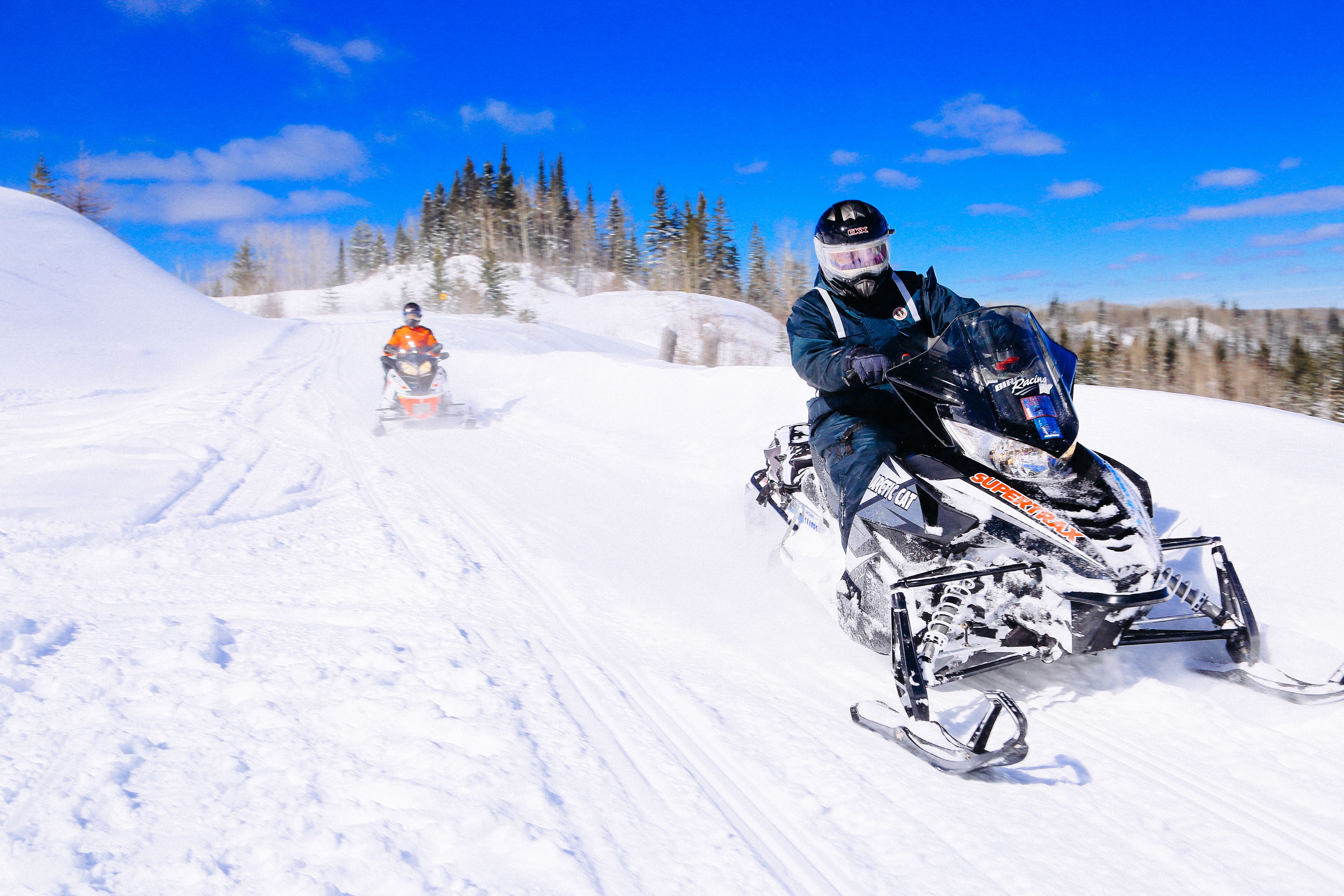 Snowmobilers ride along the crest of a snowy hill surrounded by forest on a bright sunny winter day.