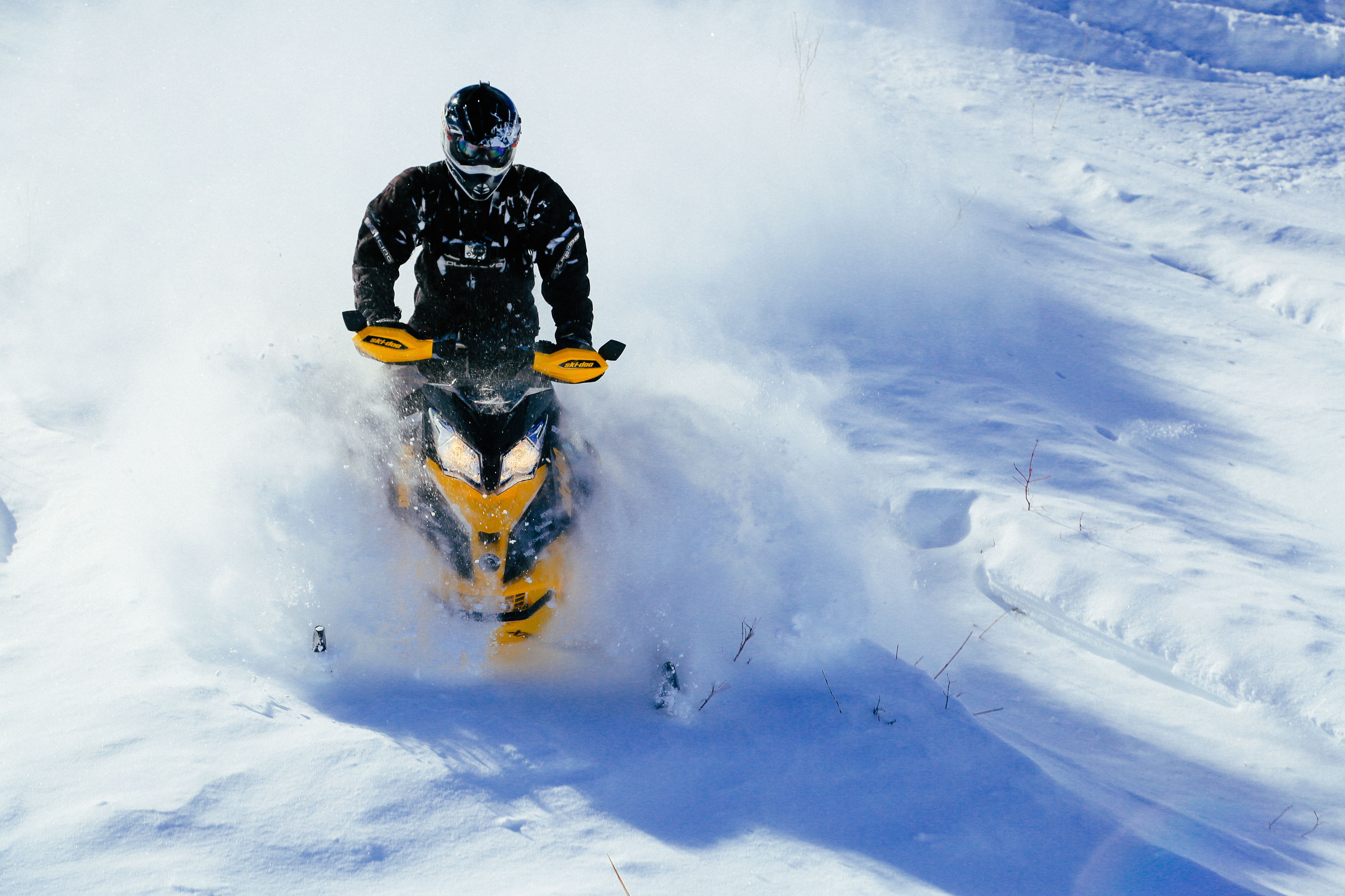 A snowmobiler rides down a powdery hill on a sunny day, kicking up a cloud of snow around them. 