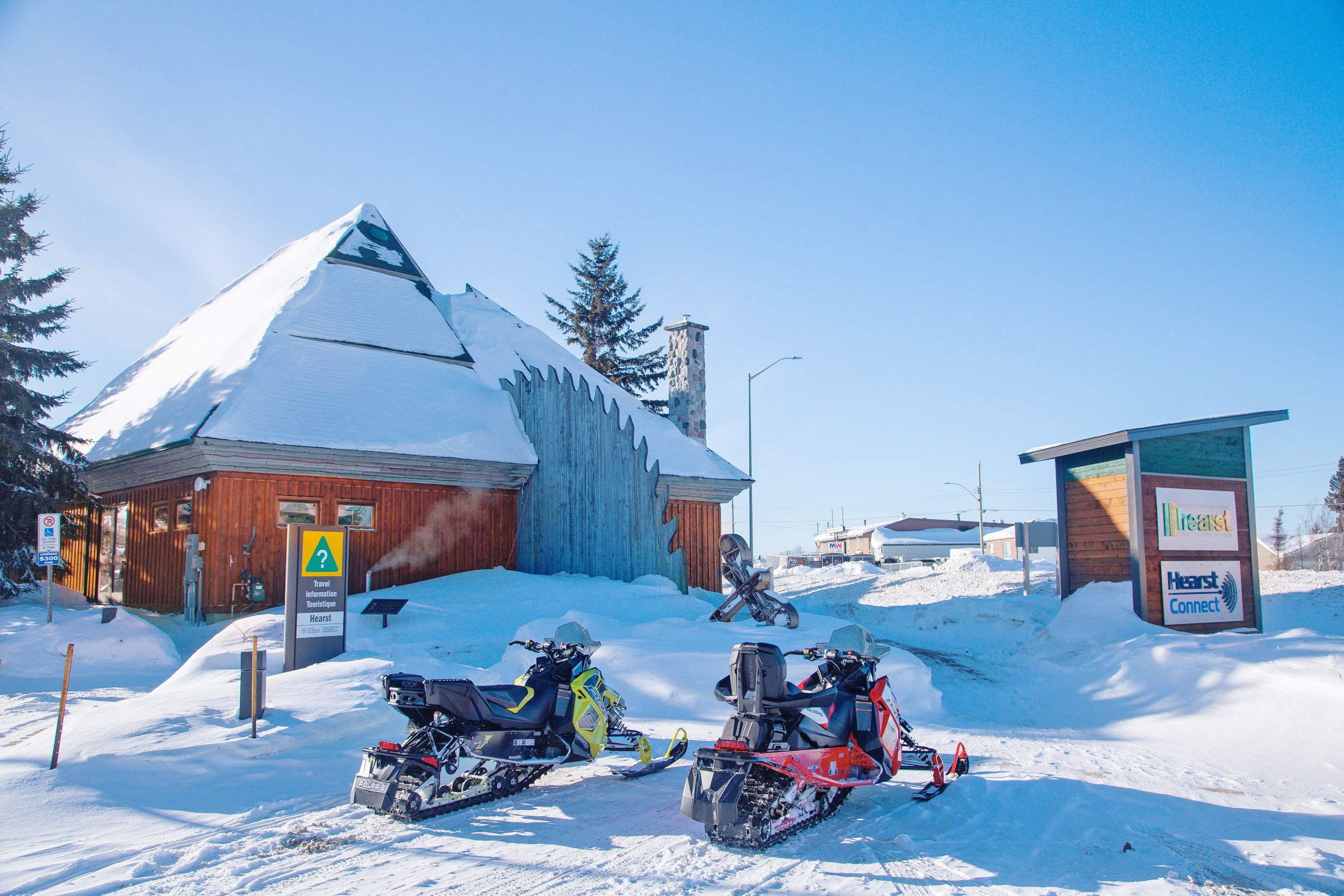 The Gilles Ganon Welcome Centre, covered in a thick layer of snow, with snowmobiles parked outside on a crisp sunny day.