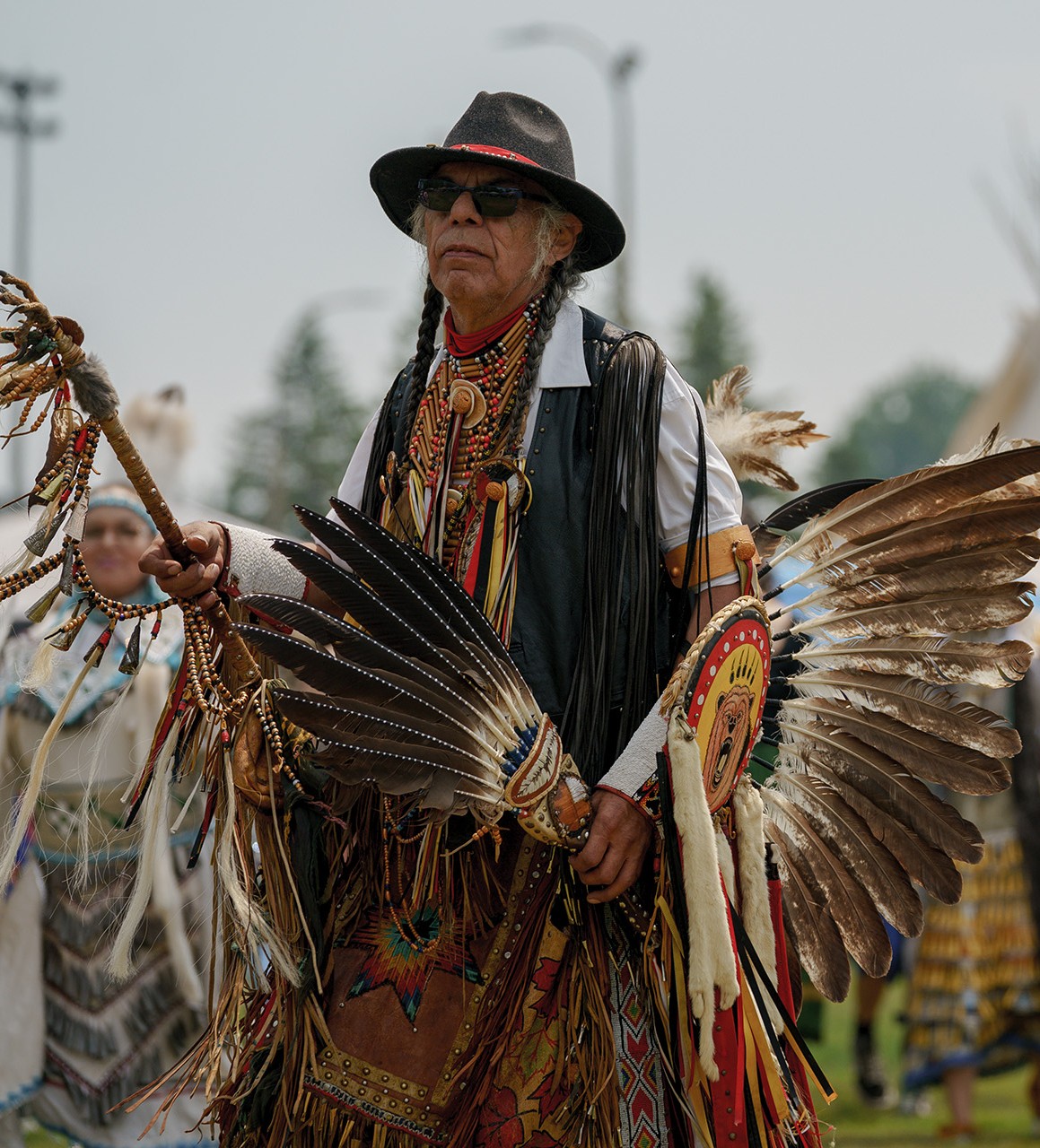 An elderly man in sunglasses, wide-brimmed hat and chicken dance regalia looks relaxed and thoughtful at a pow wow.