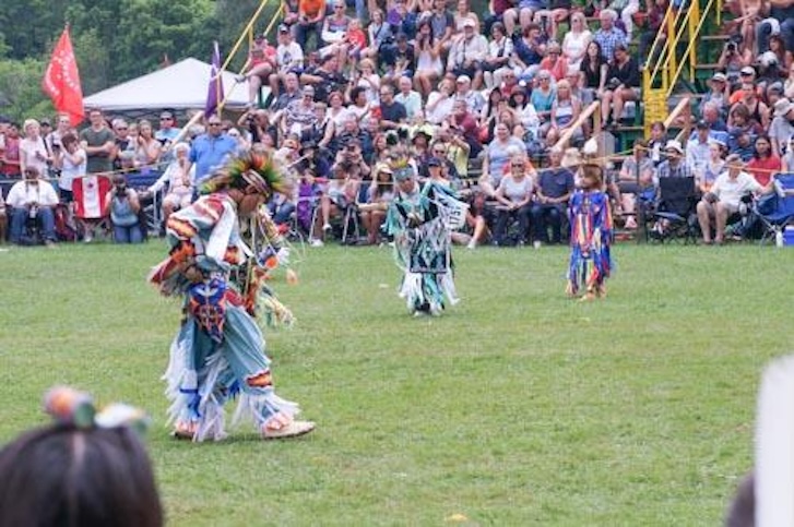 A large crowd watches from bleechers as men in regalia dance on the grass below. 