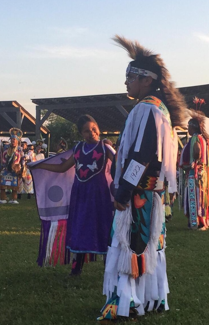 A man in regalia watches the other dancers at a pow wow at sunset, while a girl standing next to him smiles at the camera as she waits her turn to dance.