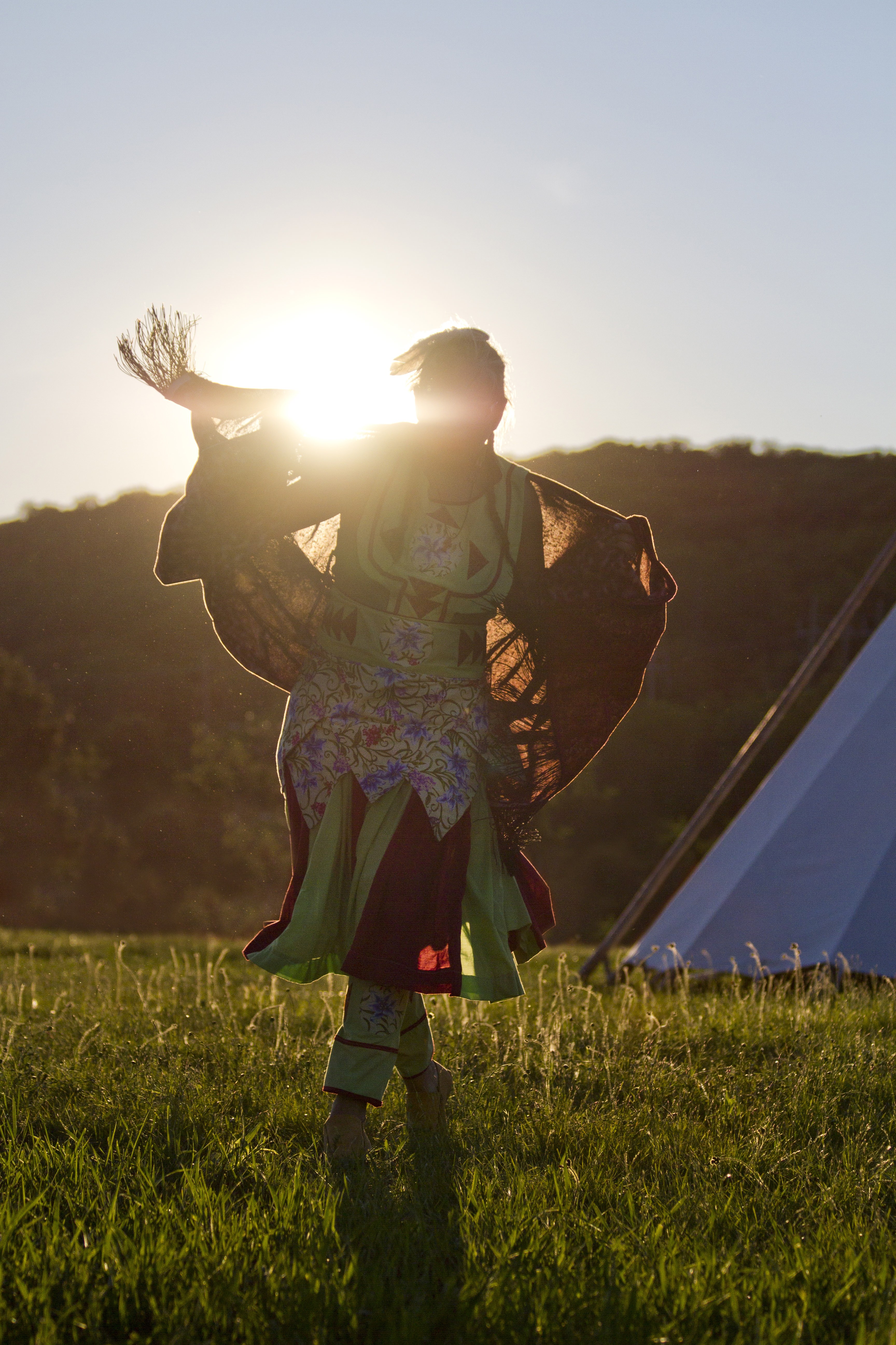 Person dancing on the grass next to a tipi at a powwow, silhouetted in the setting sun, wearing a dress and shawl.