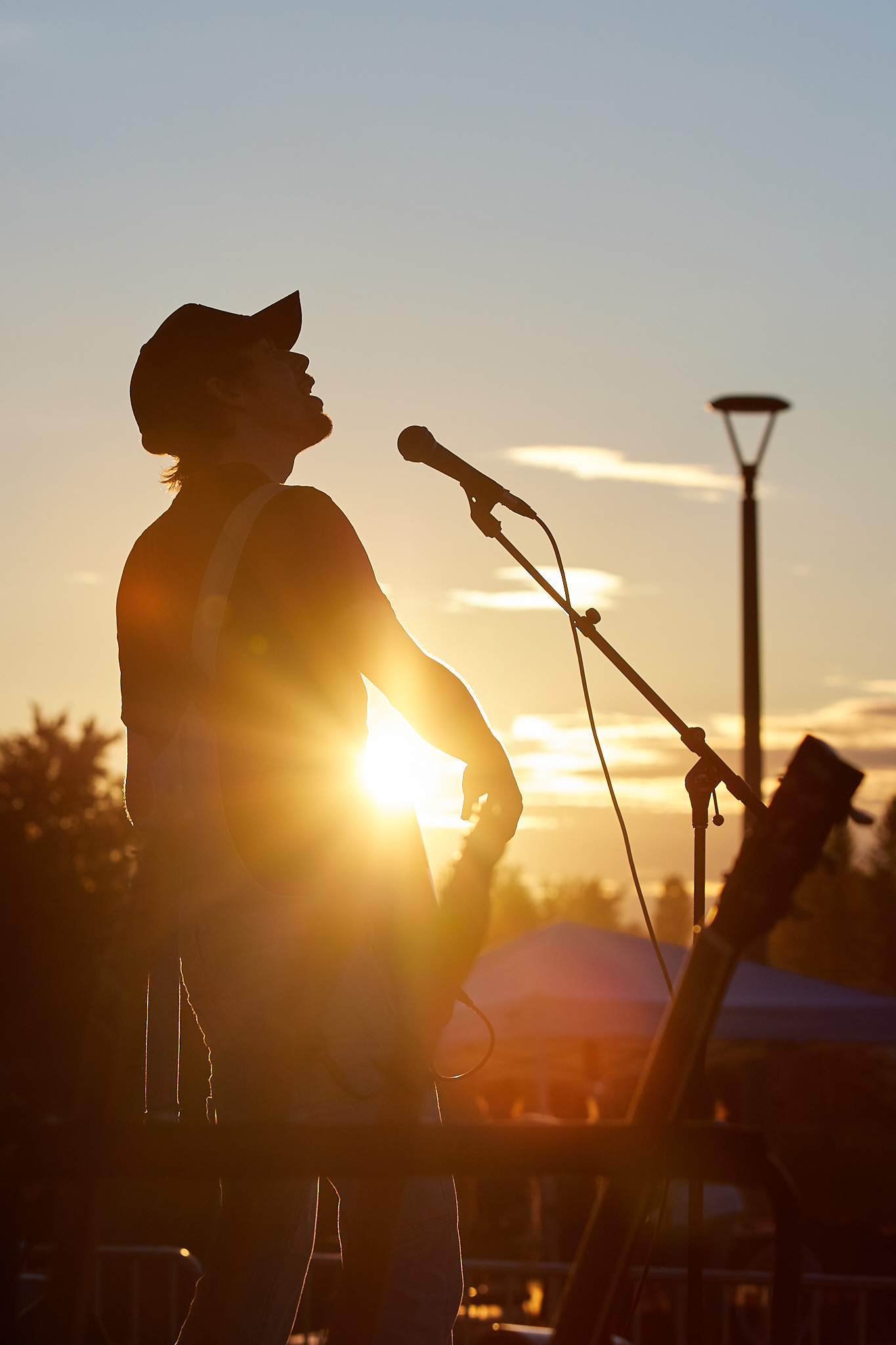 Person wearing baseball cap playing guitar and singing into a mic onstage in front of the setting sun