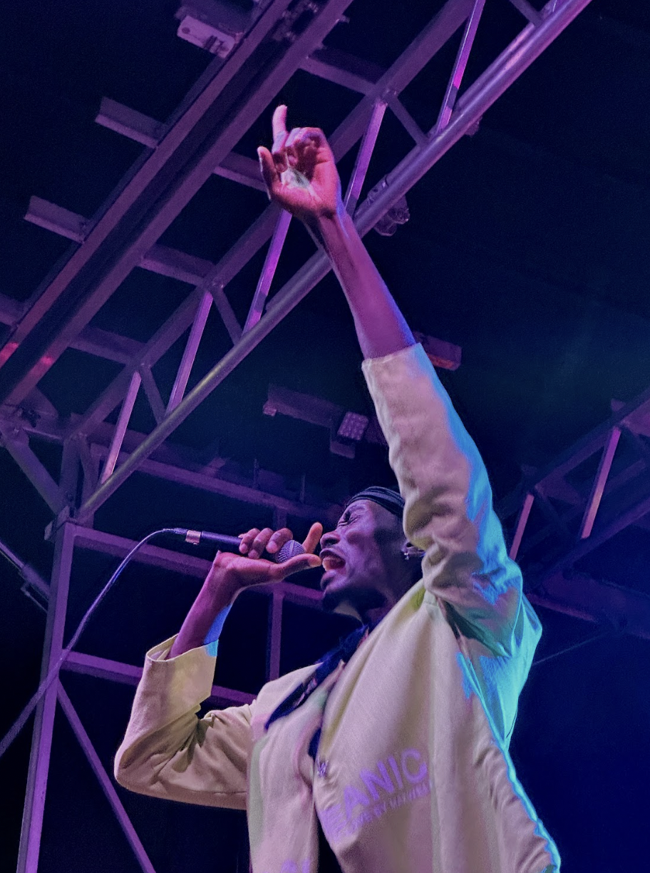 Singer Tobi on stage at Up Here wearing a black cap and white jacket pointing to the sky