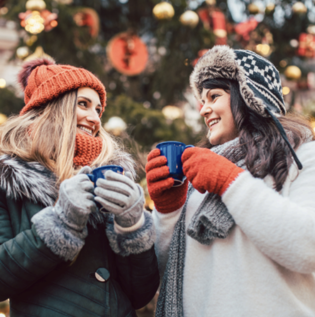 Two white women in toques drink hot chocolate in front of a decorated tree