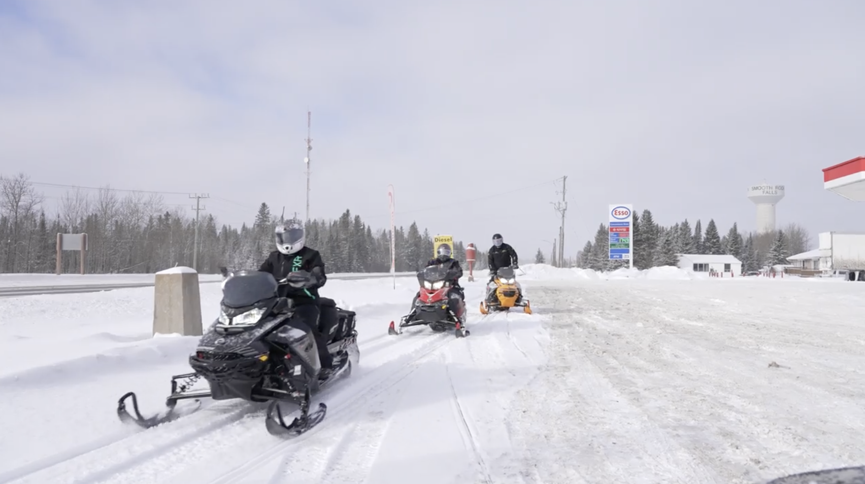 Snowmobilers ride on a snowy trail along a service road with the Smooth Rock Falls water tower in the background.