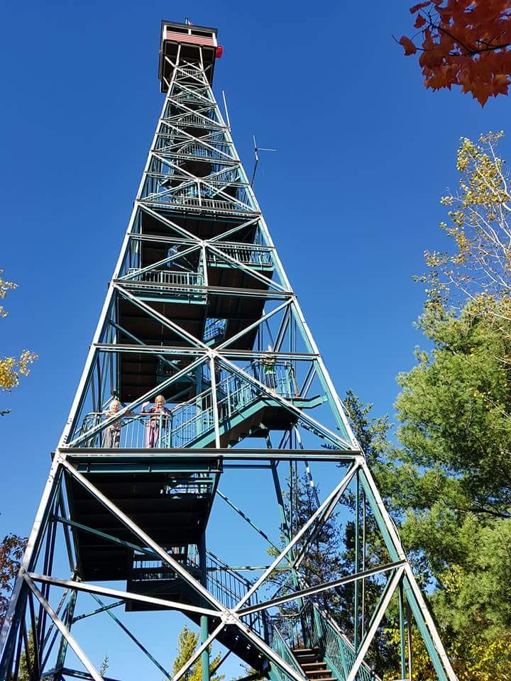 A massive fire tower with people standing stories up above the forest on a clear sunny day.