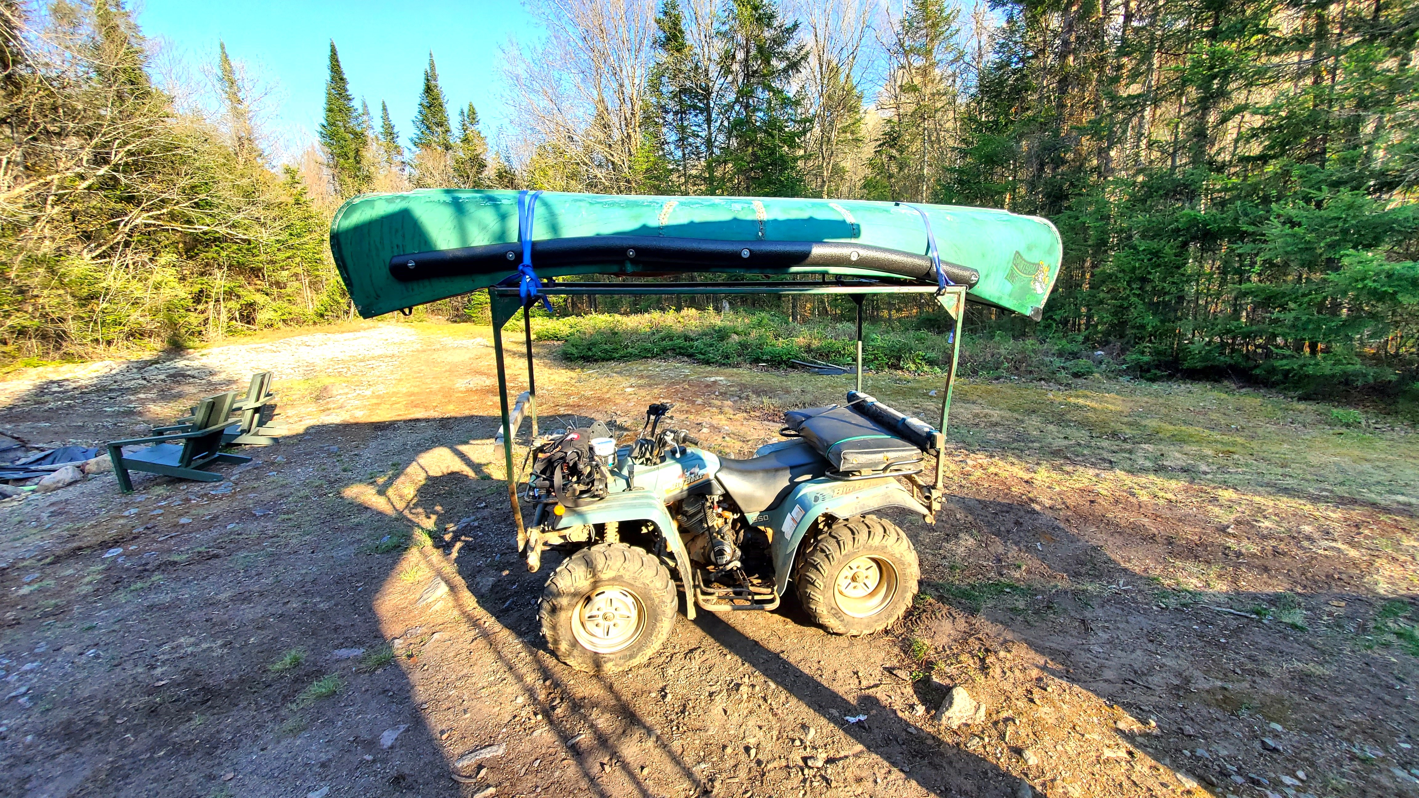 An ATV with a canoe mounted on a rack overtop sits in a clearing in the forest in the summer. 