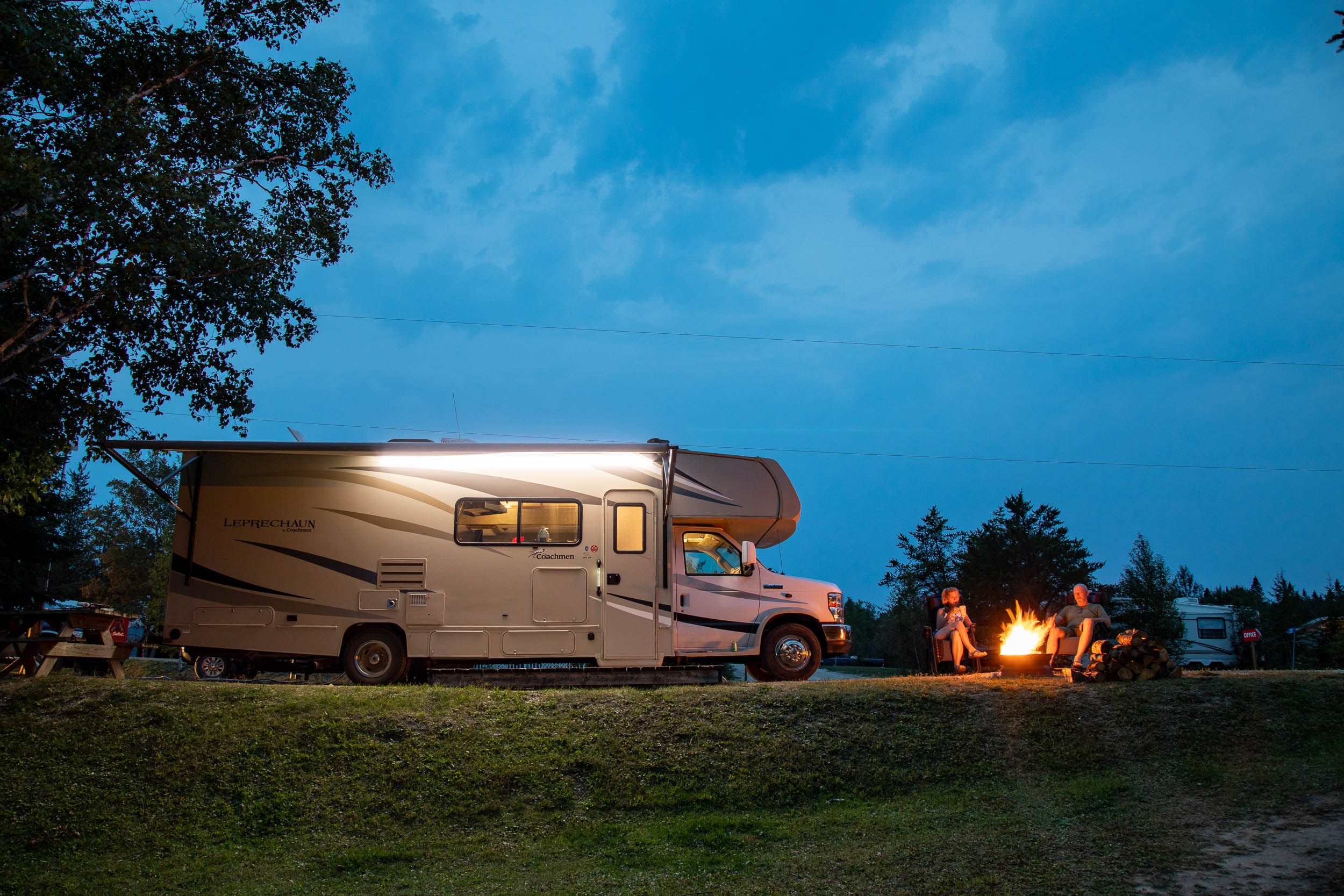 an RV parked at a treed campsite surrounded by green grass at twilight. The RV is lit up in the dark and campers sit next to it around a campfire.