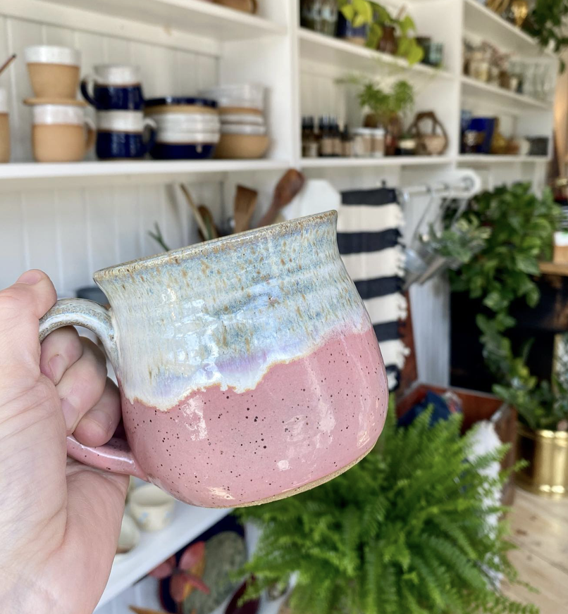 a hand holds up a handmade mug in front of shelves of other handmade pottery and green plants.