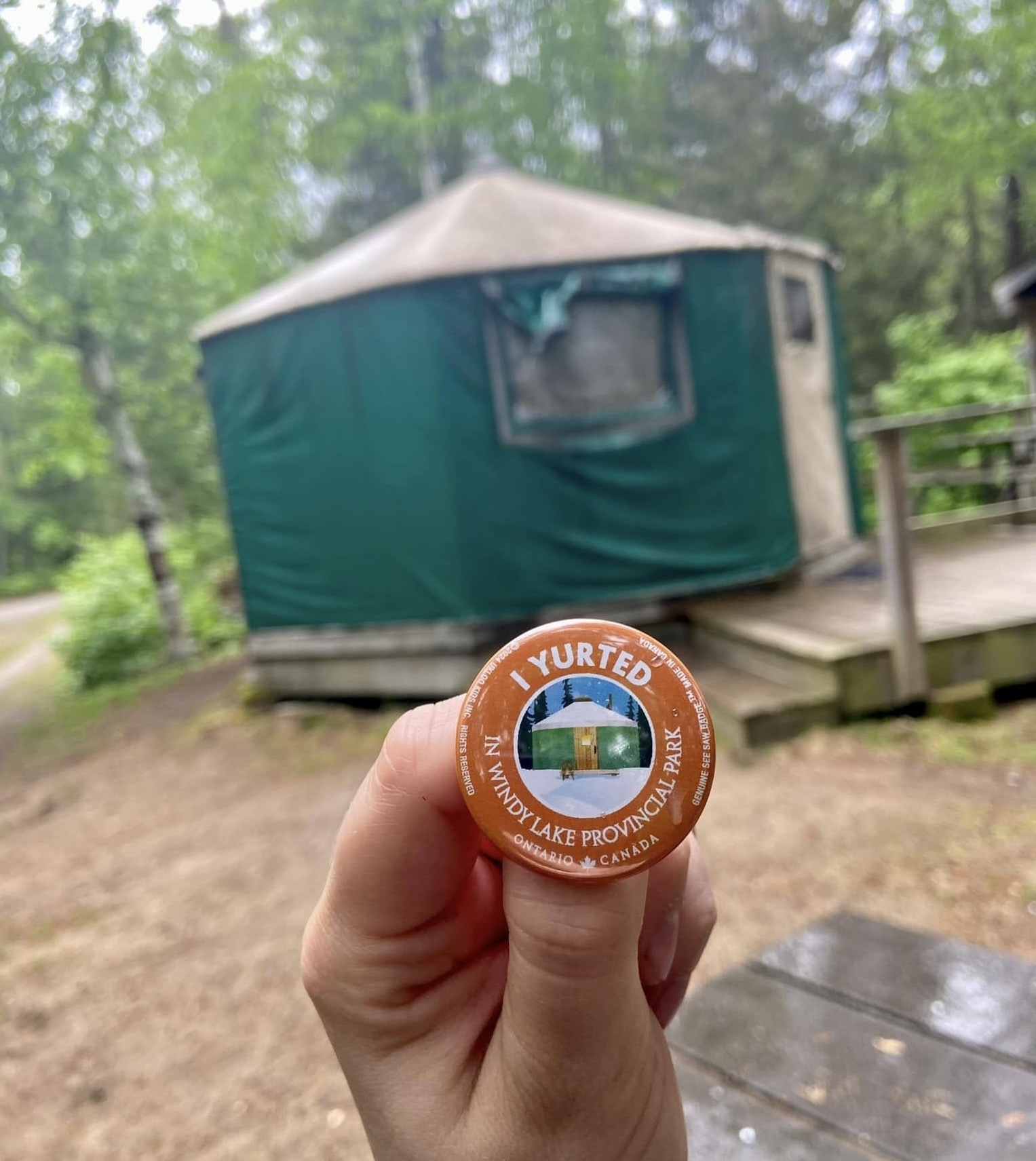 a camping yurt on a wooden platform in the forest in the summer. In the foreground a hand is holding a button that reads "I yurted in Windy Lake Provincial Park".