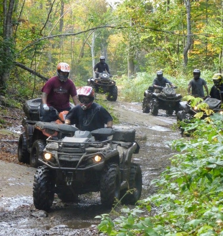 a line of ATVers ride through a muddy trail surrounded by thick green forest.