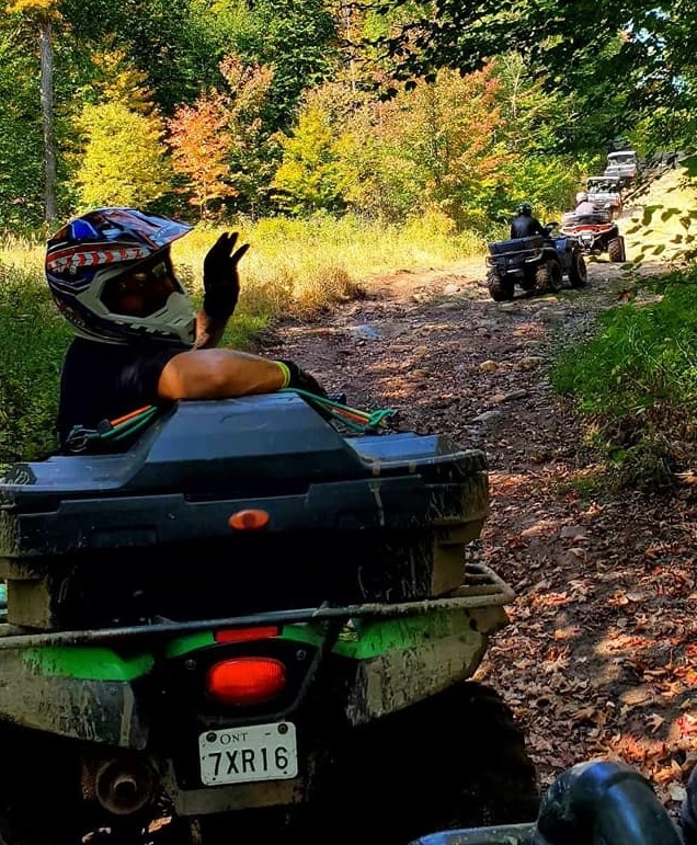 a person waving from the back of an ATV on a sunny forest trail.