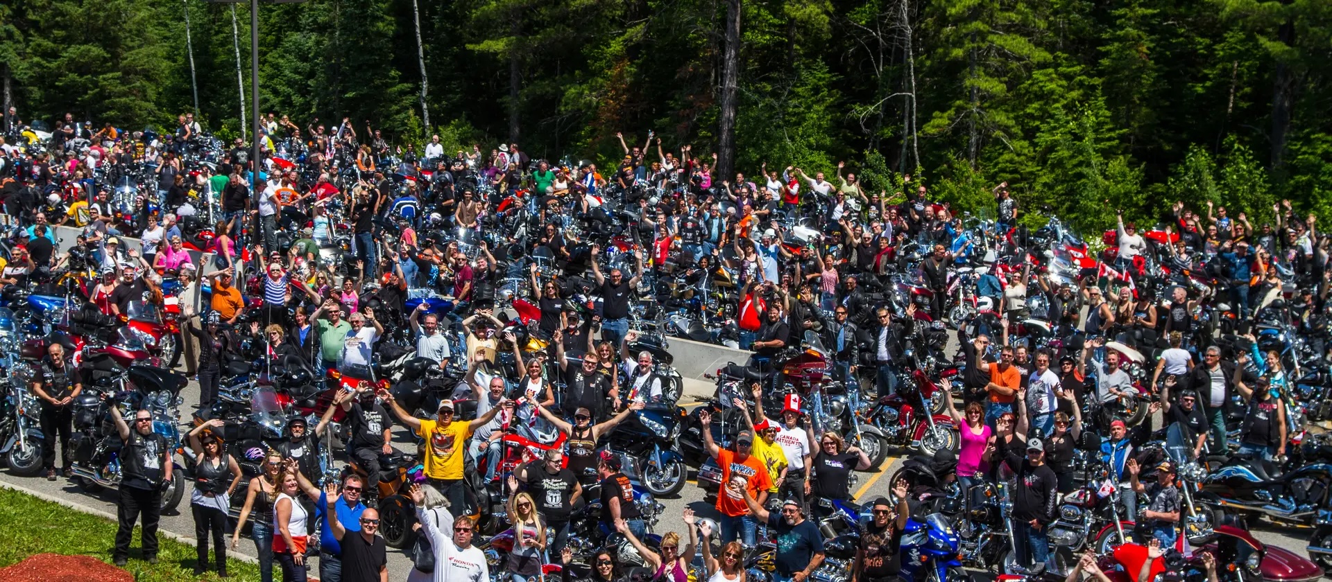 a huge crowd of smiling and cheering people with their motorcycles next to a green forest at the Bikers Reunion in New Liskeard.