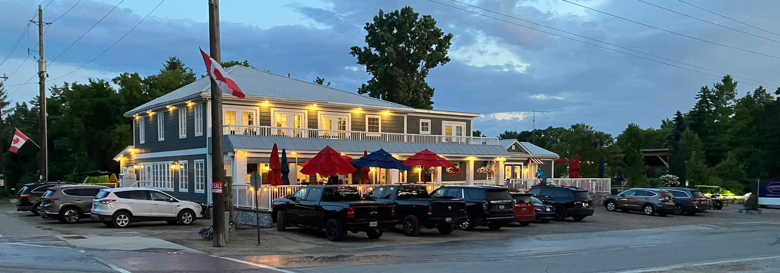 Ipperwash Beach Club; a large white historic building surrounded in green trees with red beach umbrellas on an outdoor patio, lit up on a rainy summer evening.