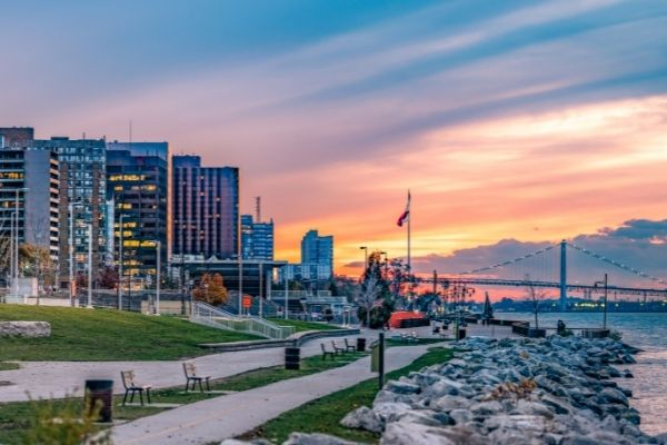 A beachfront and walkway in Windsor-Essex at sunset, next to an illuminated cityscape.